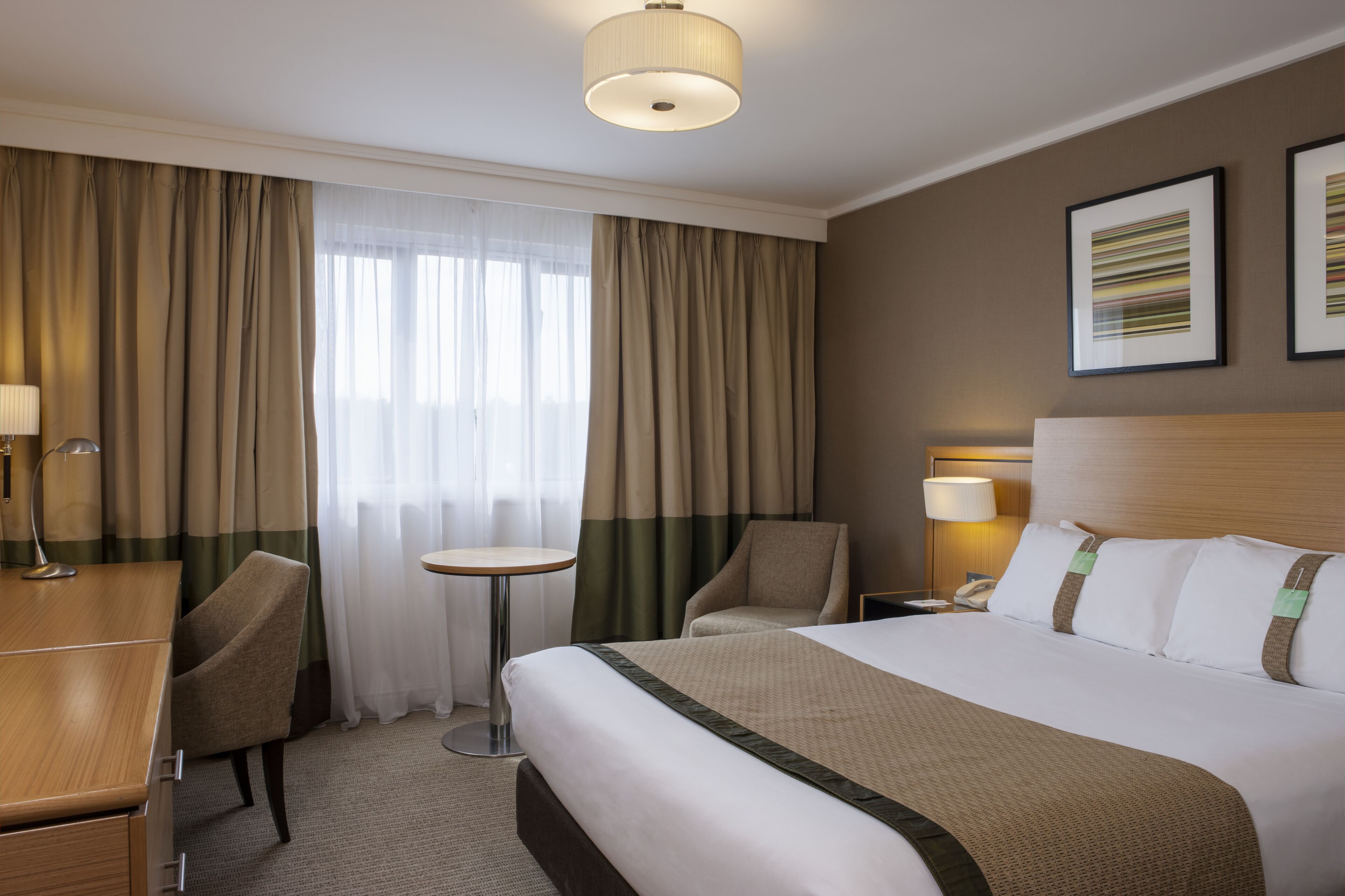 A hotel room with a bed, a desk, a chair, a round table, and a nightstand with a lamp.