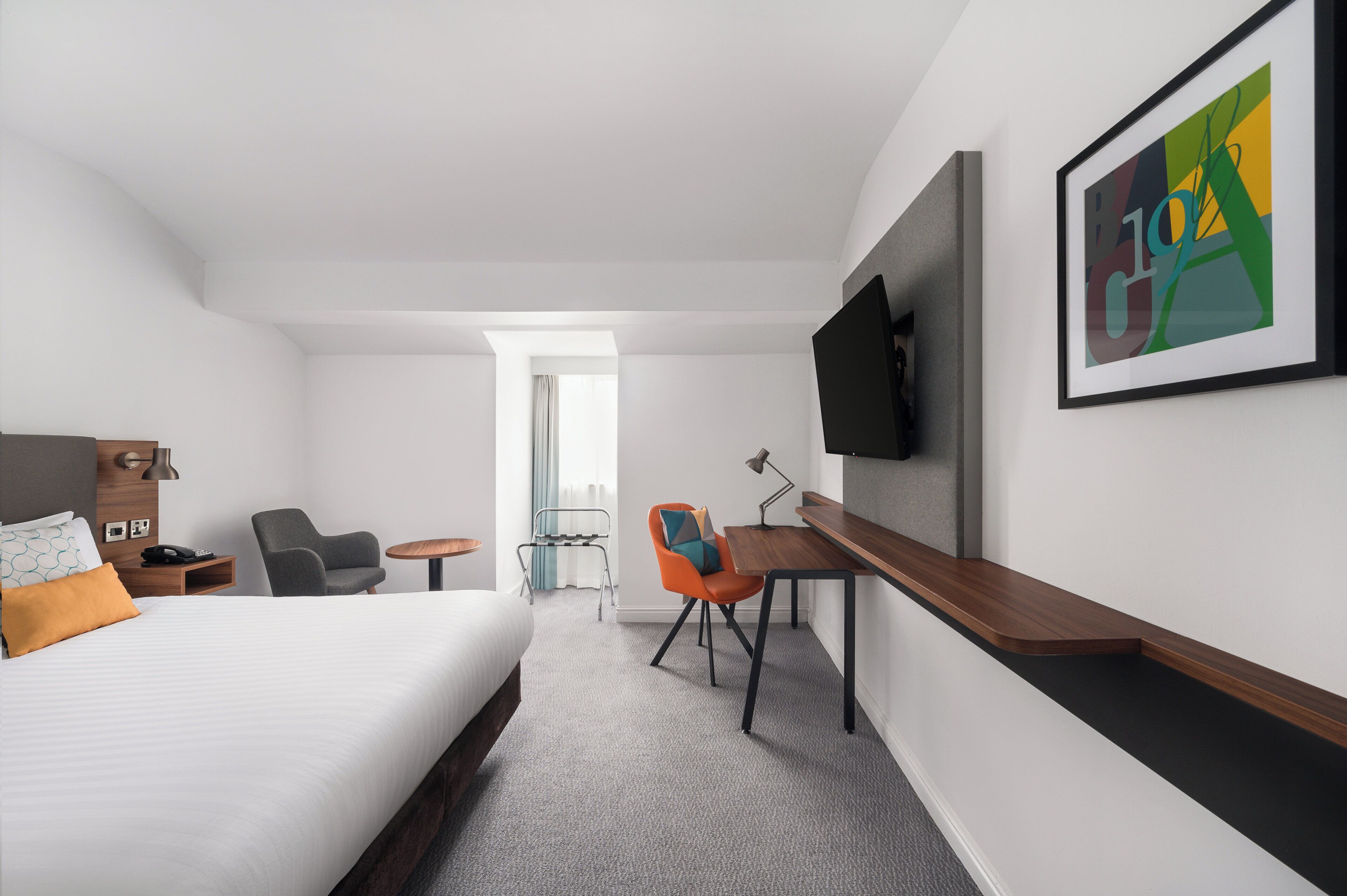 A hotel room with a bed, a desk, a chair, a TV, and a framed artwork on the wall.