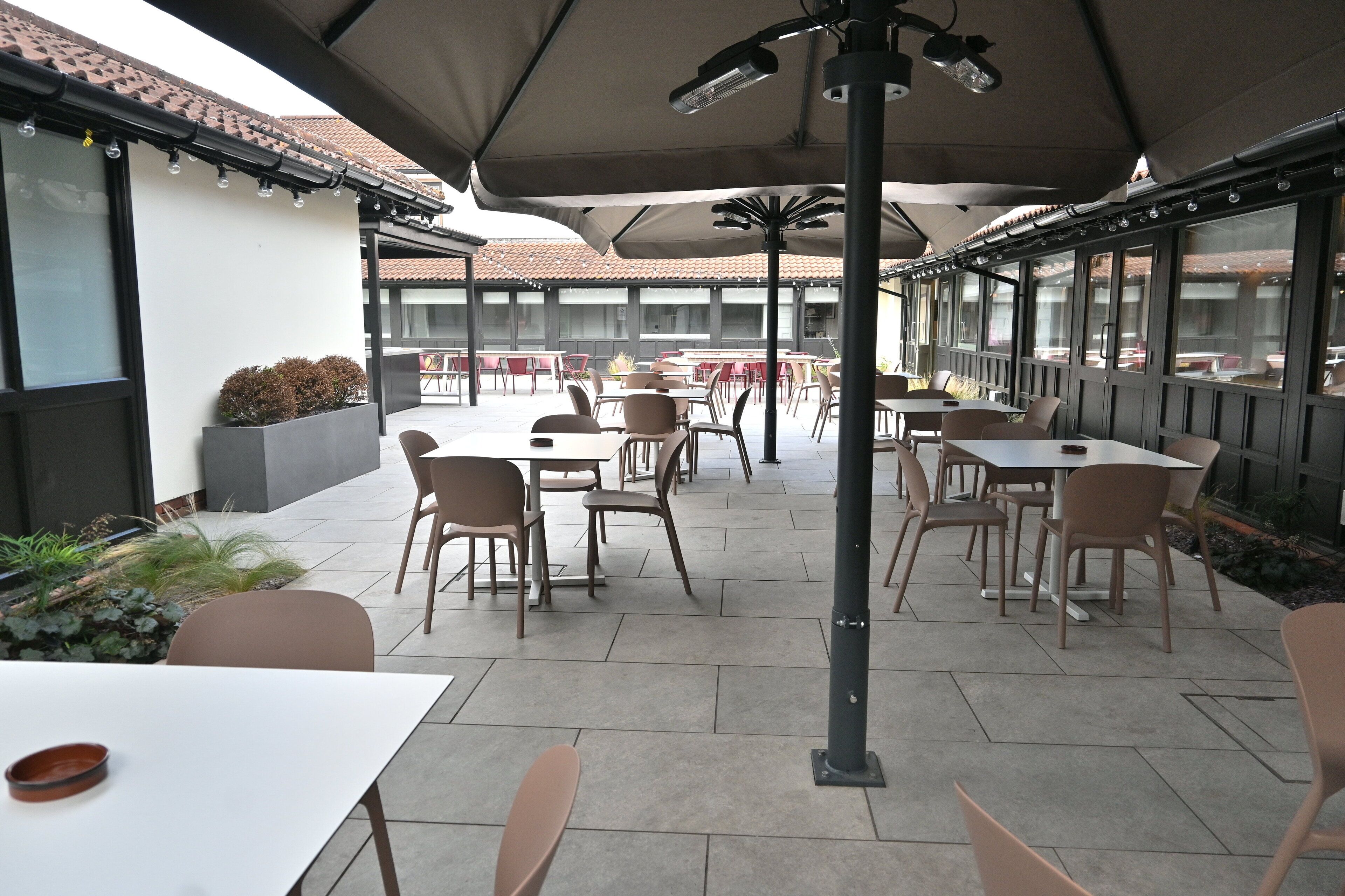 An outdoor patio area with tables and chairs, large umbrellas, and potted plants.
