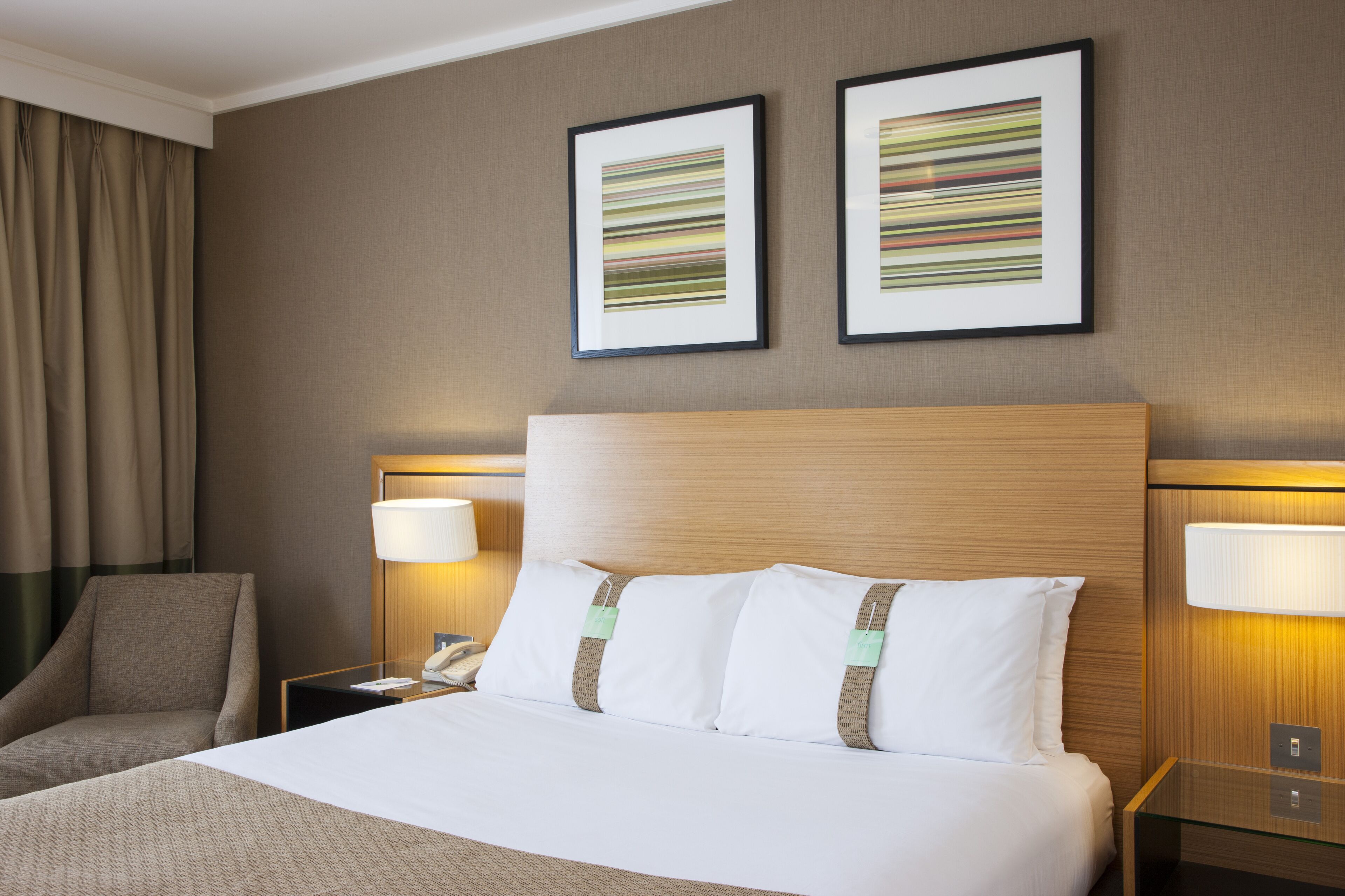 A hotel room with a large bed, two bedside tables with lamps, a chair, and two framed artworks on the wall.