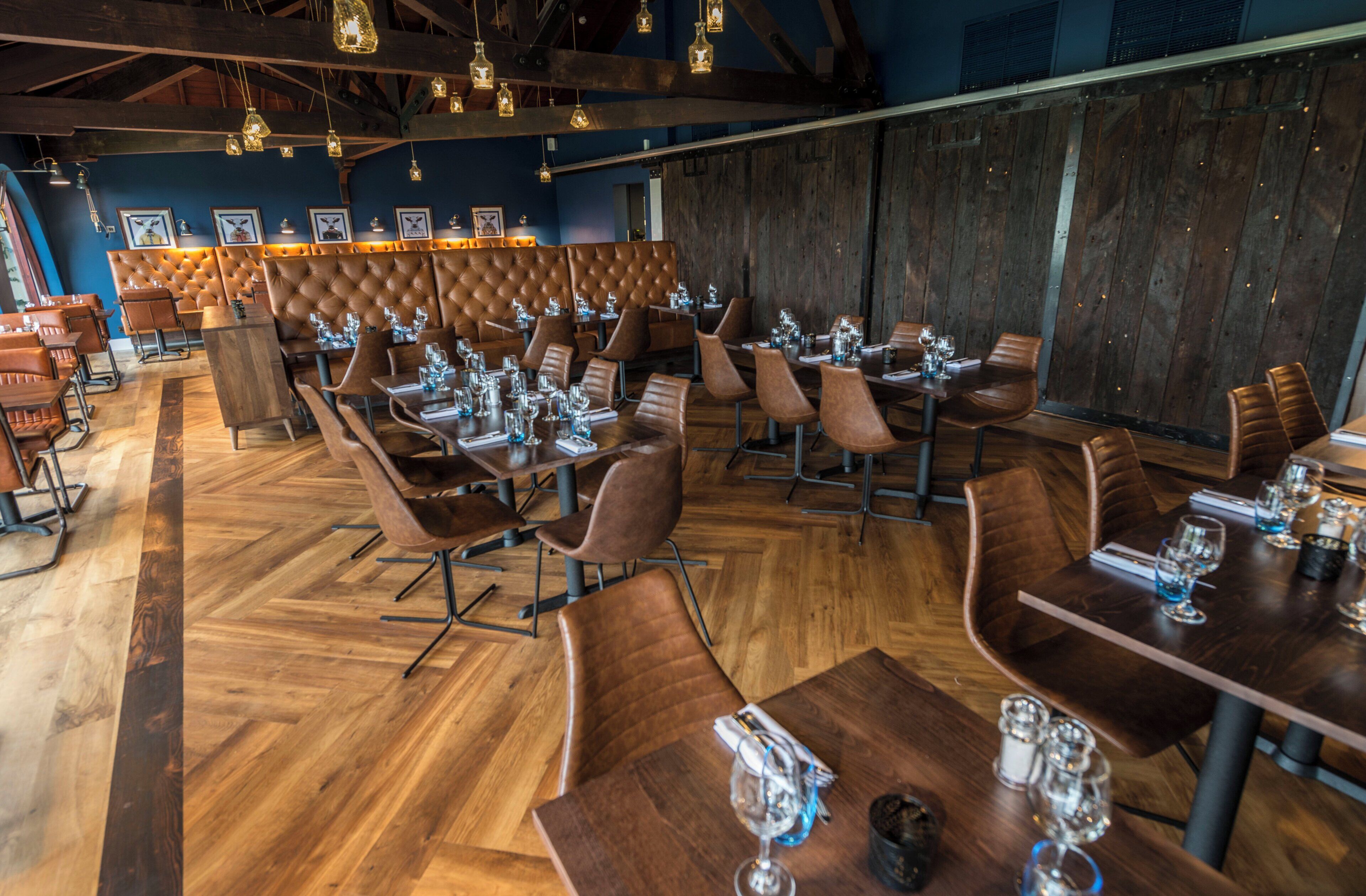 L’intérieur d’un restaurant avec des tables et des chaises en bois, un plafond haut orné de luminaires suspendus et un mur bleu décoré de tableaux encadrés.