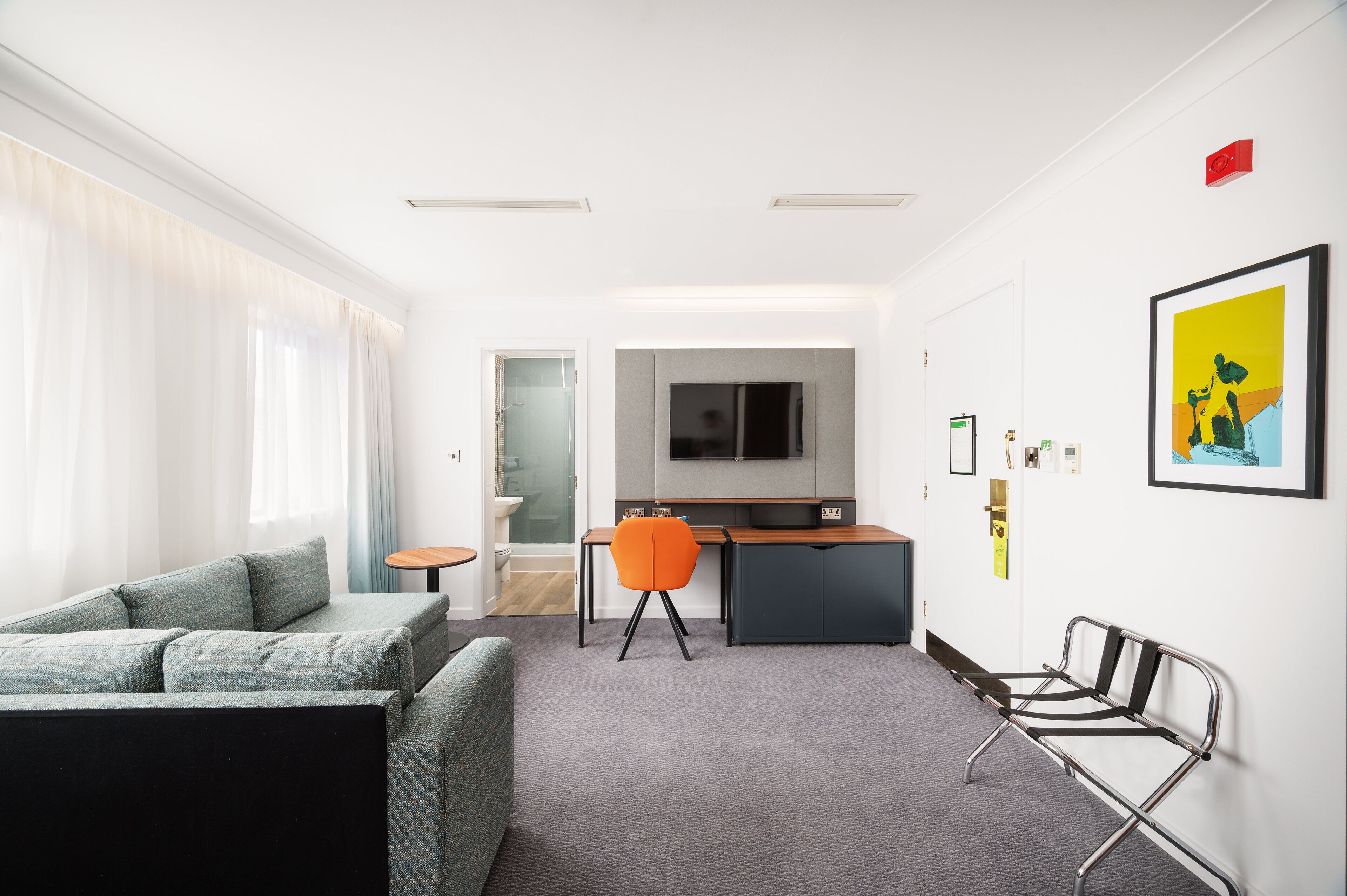 A modern hotel room with a grey sofa, a small round table, a desk with a chair, a TV, and a framed picture on the wall.