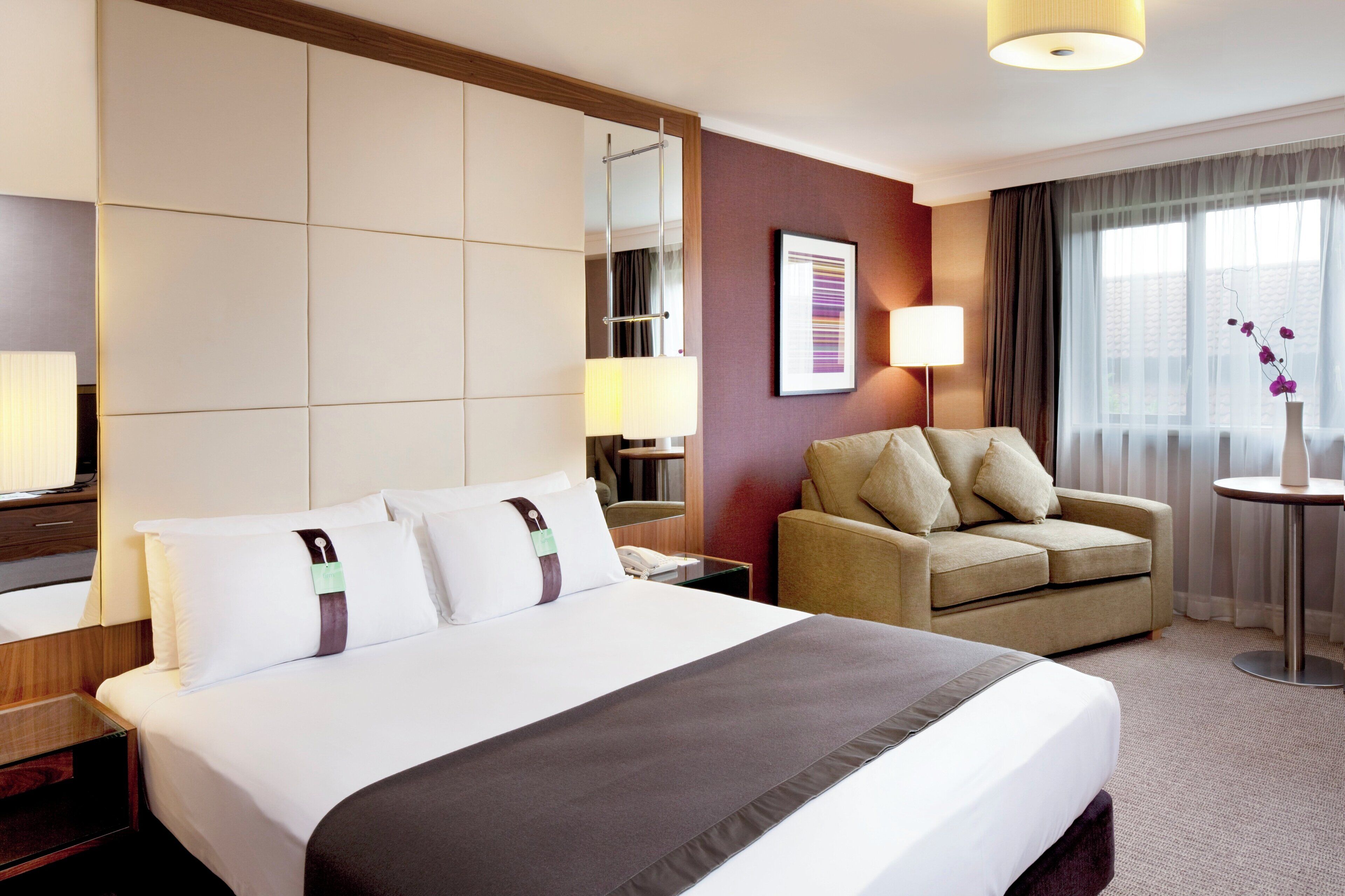 Habitación de hotel con una cama grande, un sofá, una mesita, una lámpara, un espejo y una ventana con cortinas.