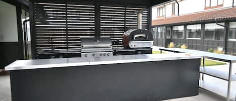 Un área de cocina exterior con una parrilla empotrada, un horno de pizza y una encimera de acero inoxidable.