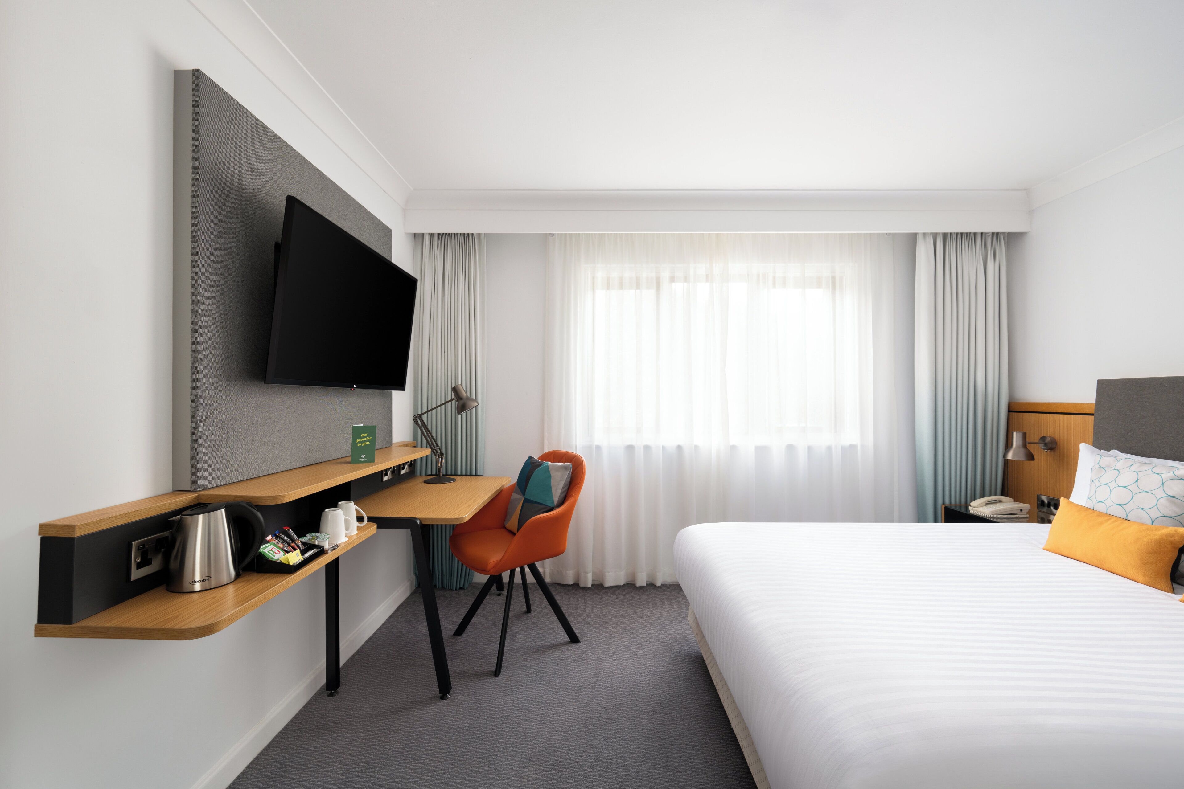 A hotel room with a bed, a desk with a chair, a flat-screen TV mounted on the wall, a coffee maker, and a telephone on the nightstand.