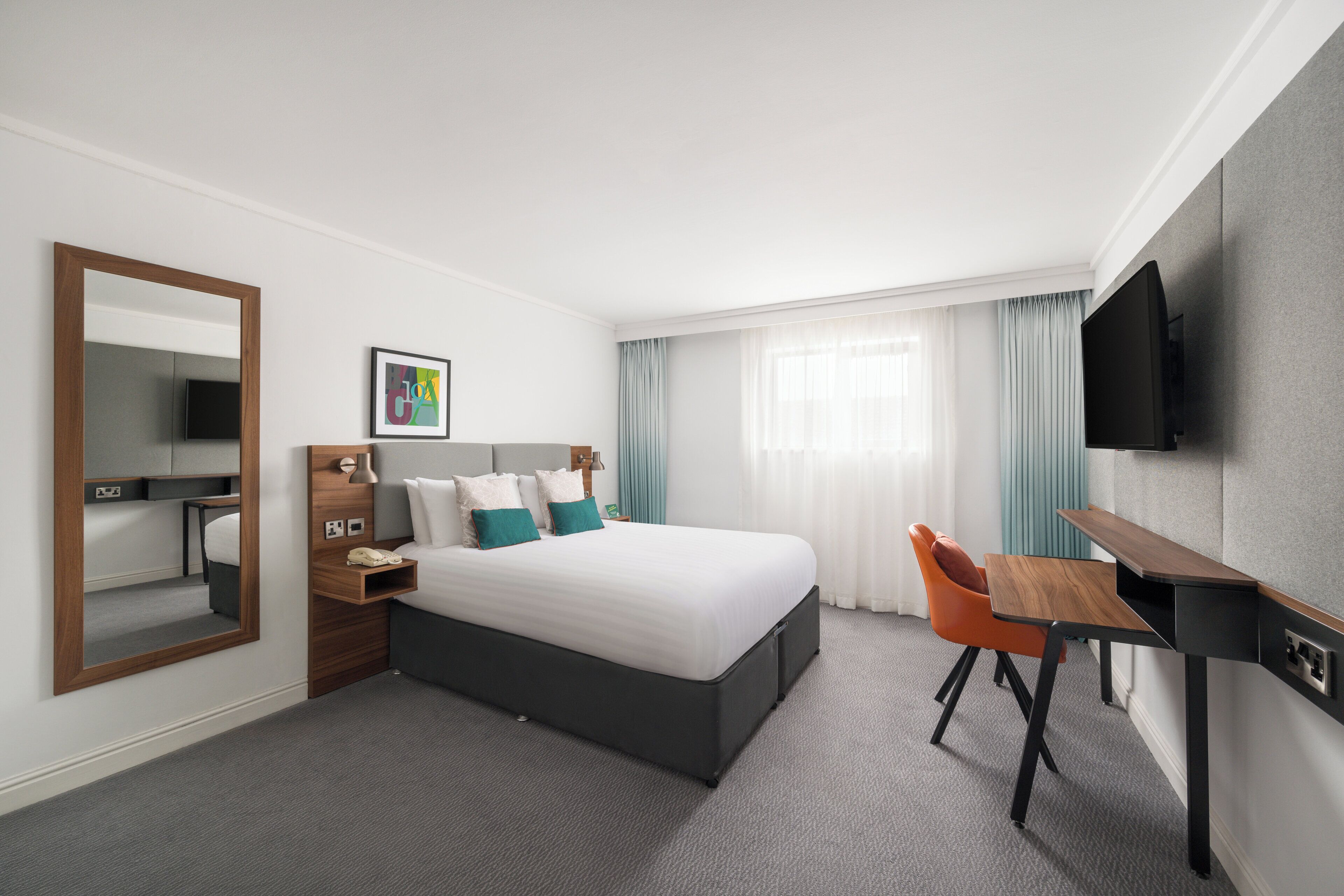 A hotel room with a bed, a desk, a chair, a TV, and a mirror.