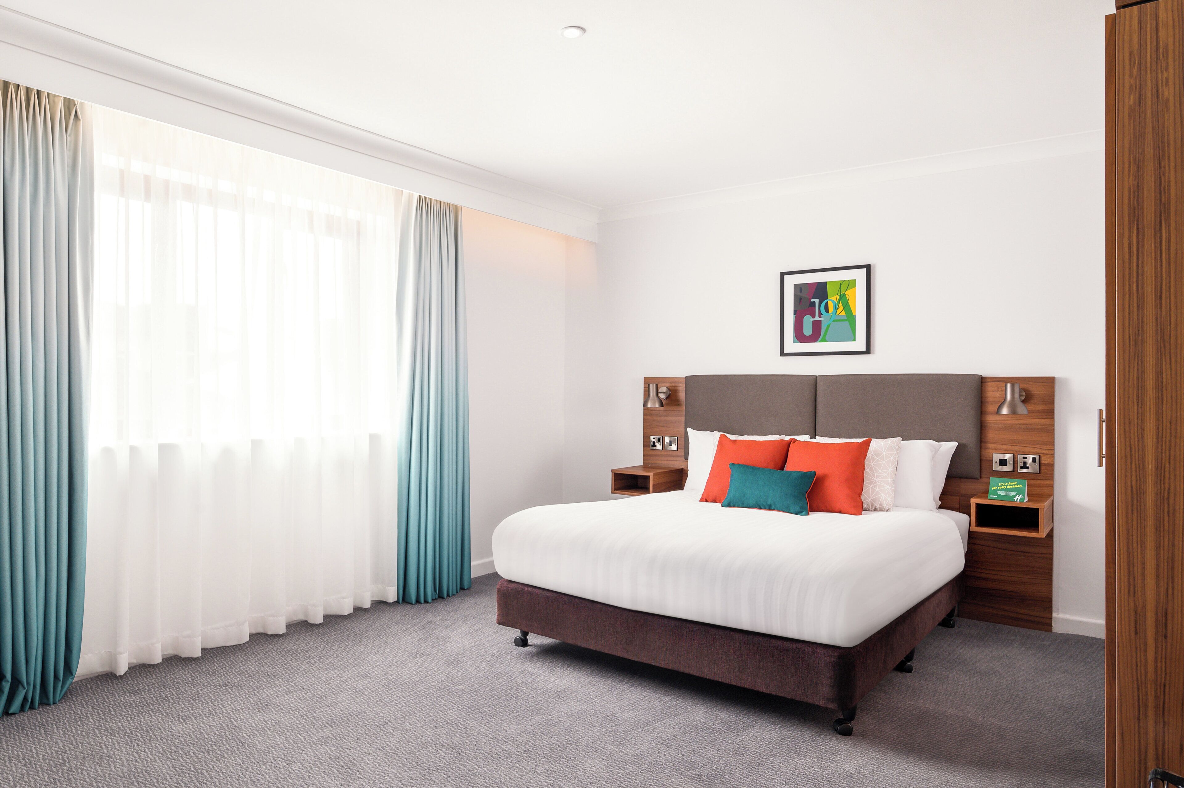 A hotel room with a large bed, wooden headboard, two bedside tables, a nightstand, a framed picture on the wall, and a window with curtains.