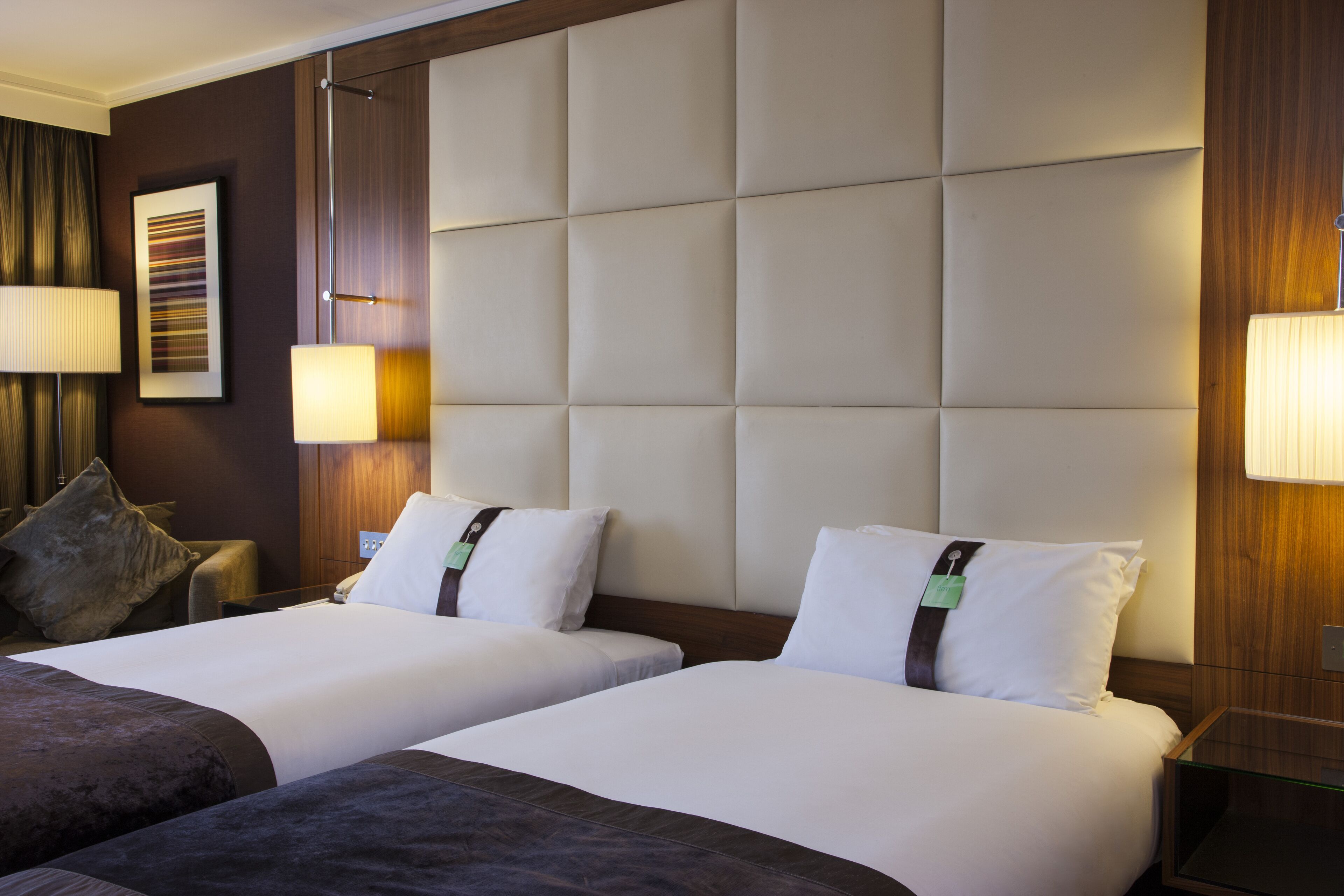 A hotel room with two beds, a large padded headboard, bedside lamps, and a framed picture on the wall.