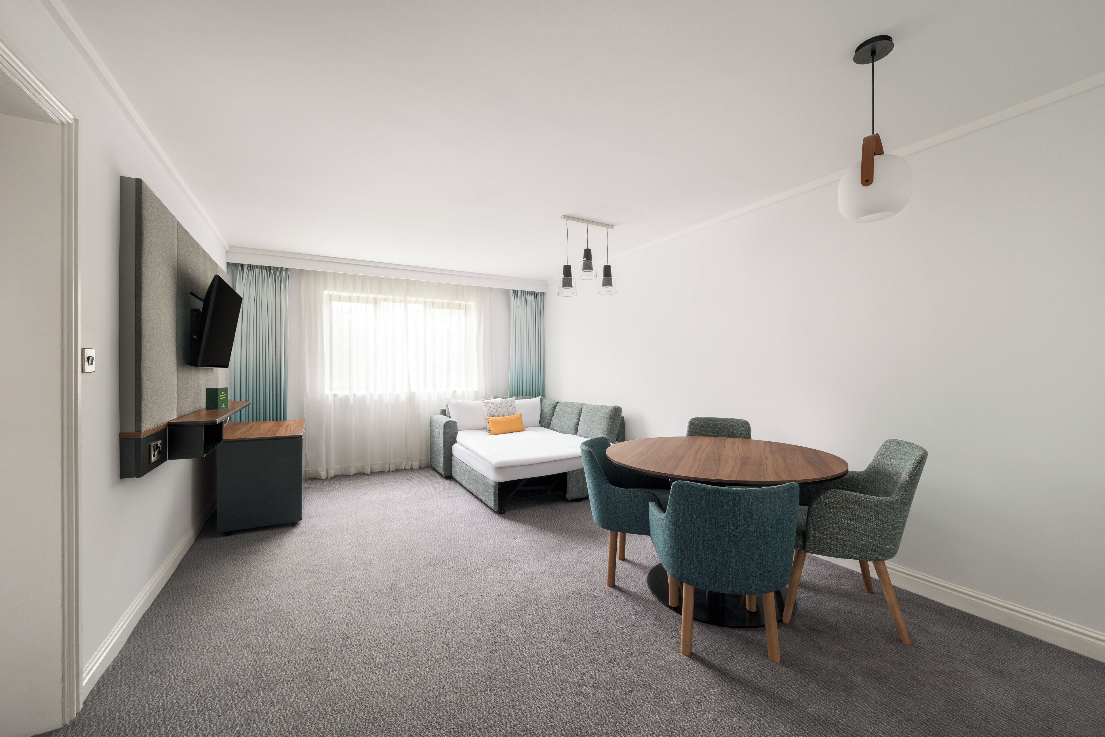 A modern hotel room with a sofa bed, a dining table with chairs, a TV mounted on the wall, and a ceiling light fixture.