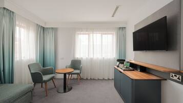 A modern hotel room with a sofa, two armchairs, a small round table, a flat-screen TV mounted on the wall, and a wooden side table with a coffee machine.