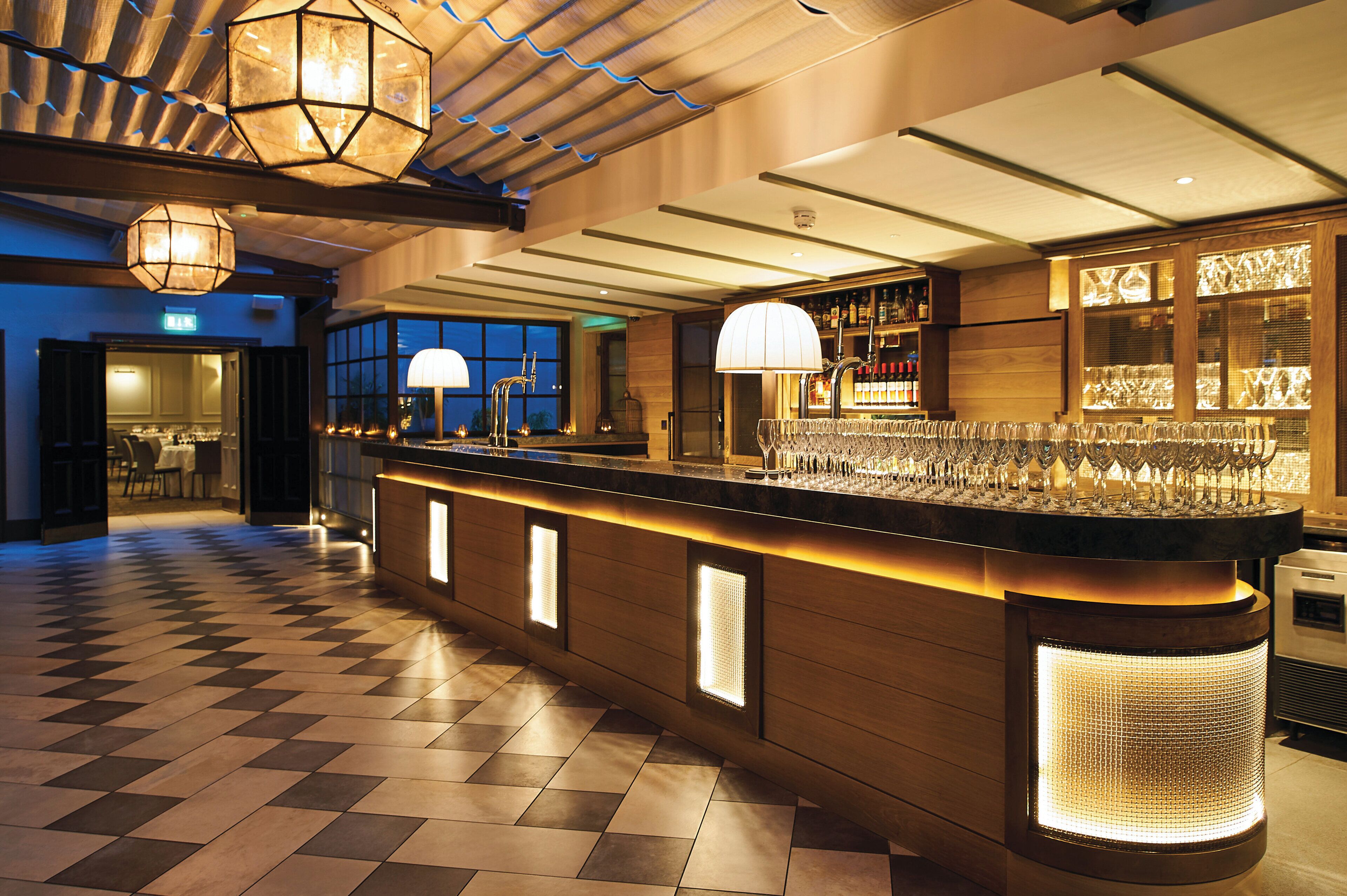 A bar area with a checkered floor, bar counter with a lighted base, and overhead lighting fixtures.