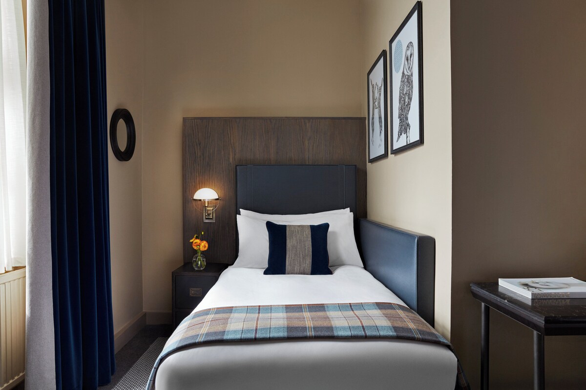 A hotel room with a bed, a nightstand with a lamp, a framed picture, and a small table with a book.