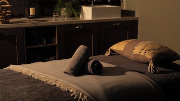 A neatly made bed with a blanket and two pillows, a bedside table with a glass and a candle, and a kitchen area with a sink and various items on the counter.