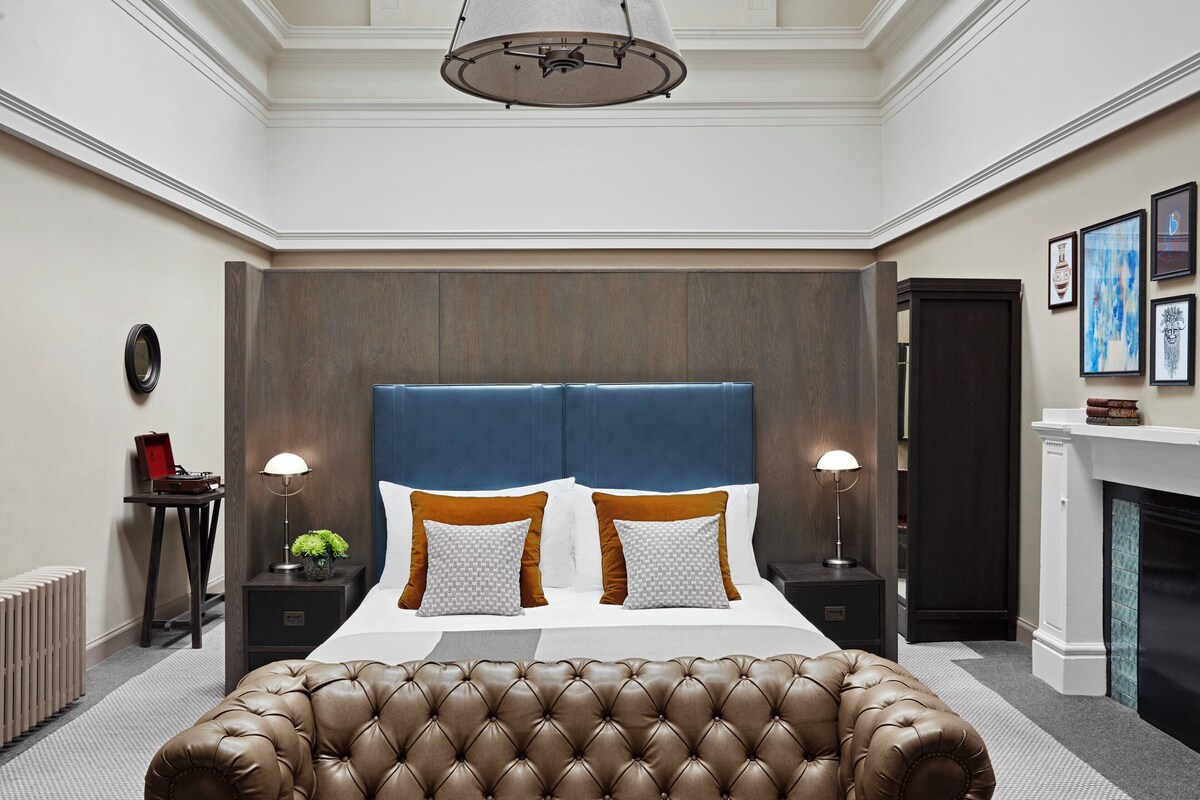 A hotel room with a large bed, a tufted headboard, two nightstands, a lamp, a fireplace, and a mirror on the wall.