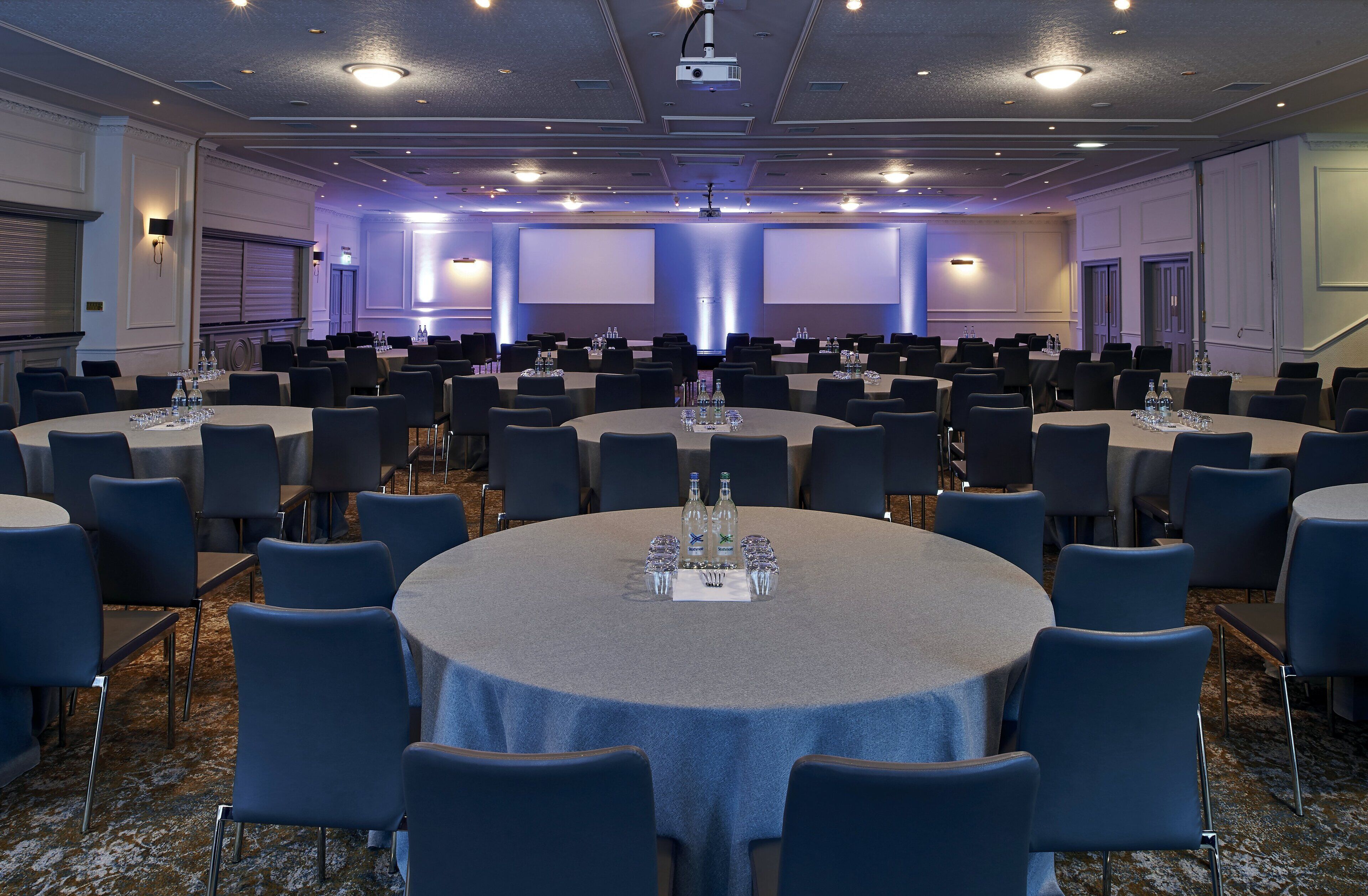 Une salle de conférence équipée de plusieurs tables rondes, chacune étant pourvue de chaises, de bouteilles d’eau et de verres.