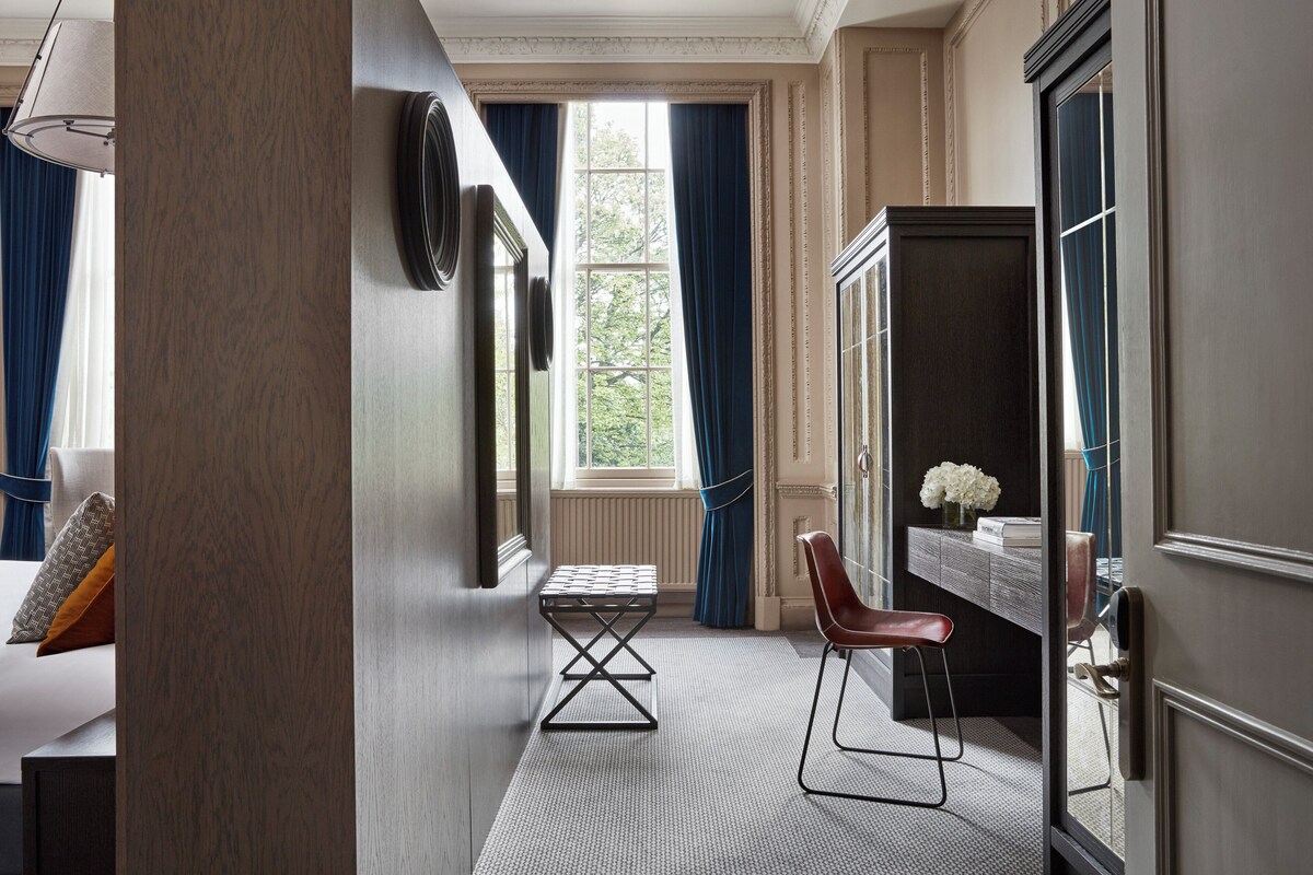 A hotel room with a large window, a desk with a chair, and a bed with pillows.