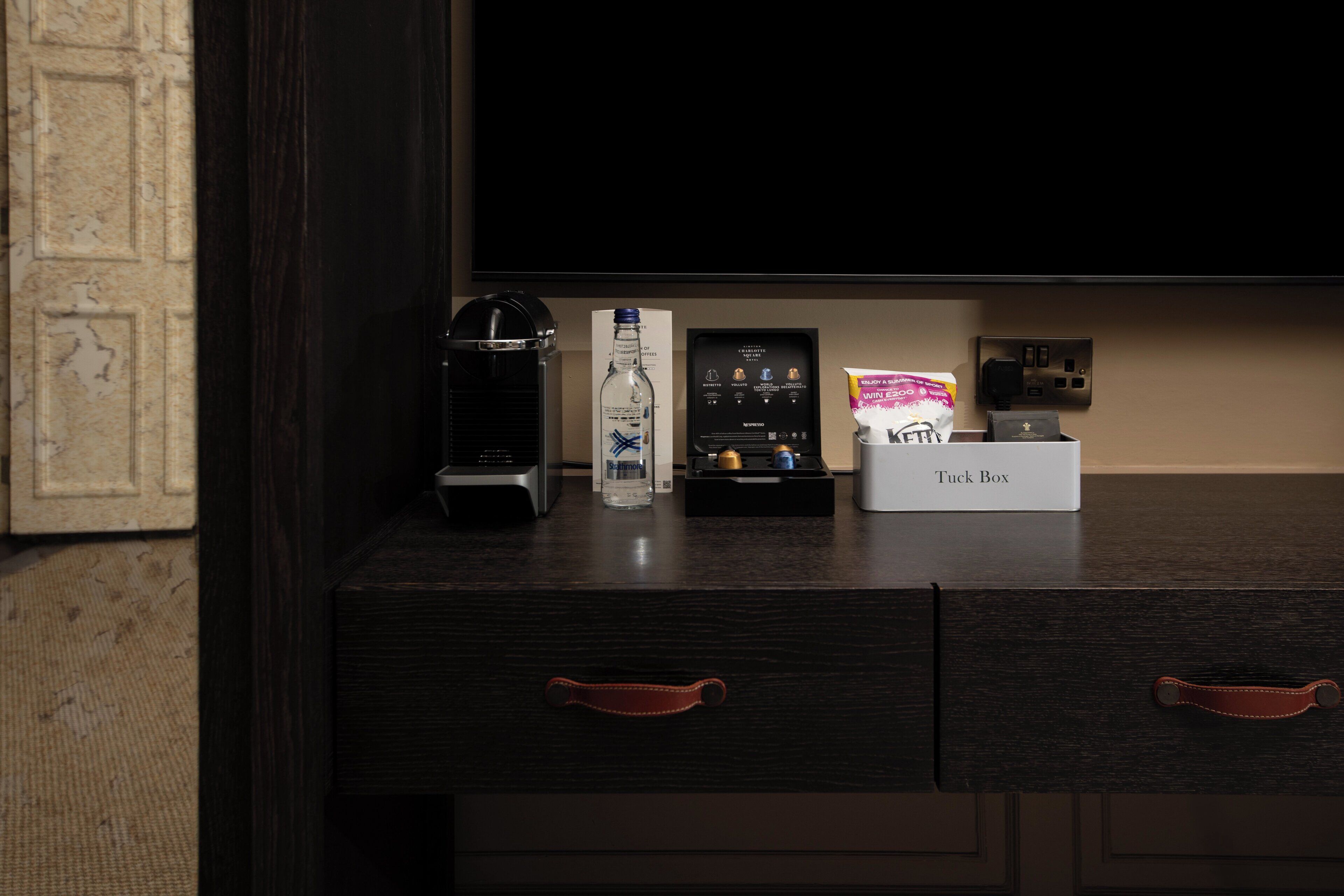 A dark wooden desk with a Tuck Box, a water bottle, a coffee maker, a framed certificate, and a small box of snacks.