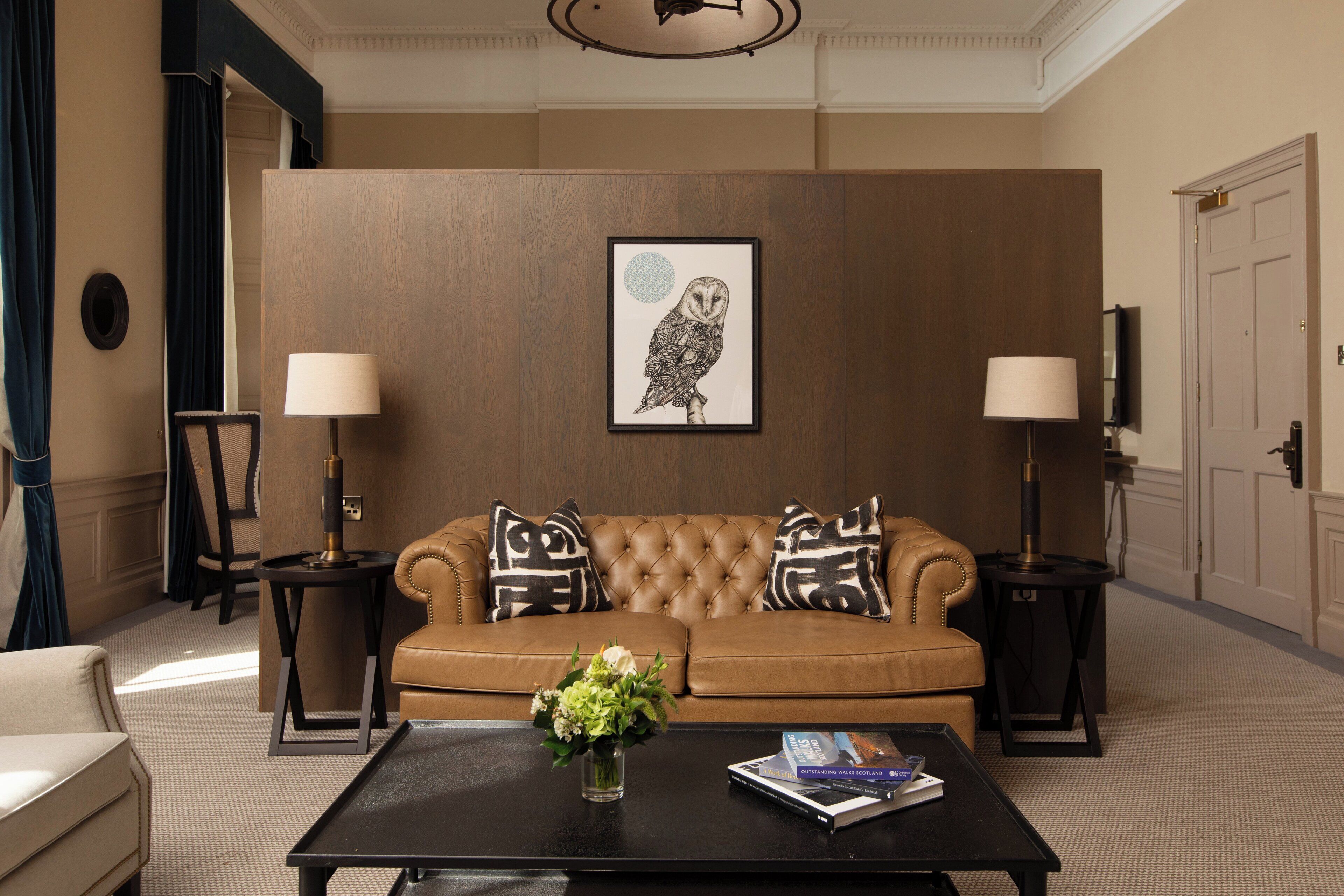 A room with a brown leather sofa, two side tables with lamps, a framed owl artwork on the wall, and a coffee table with a vase and books.