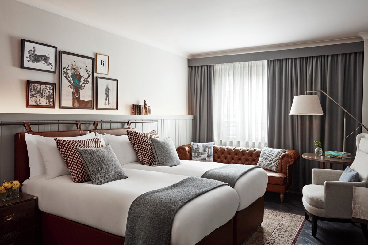 A hotel room with two beds, a sitting area with a chair, a lamp, and framed artwork on the wall.