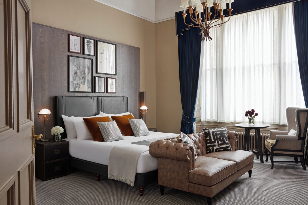 A hotel room with a large bed, a sofa, a chair, a table, and a chandelier.