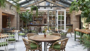 A spacious indoor patio with a high ceiling, featuring a central round table surrounded by wicker chairs, a bar area with shelves of bottles, and various potted plants. - Kimpton Charlotte Square by IHG (Edinburgh)