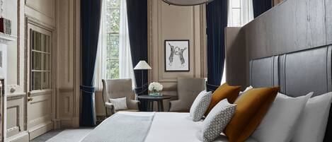 A hotel room with a large bed, a round table, two chairs, a lamp, and a large window with curtains.