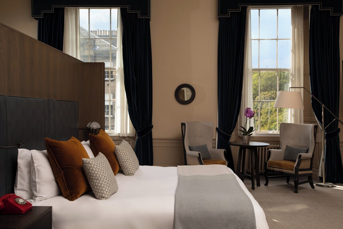 A hotel room with a large bed, two armchairs, a small table, a lamp, and a telephone.