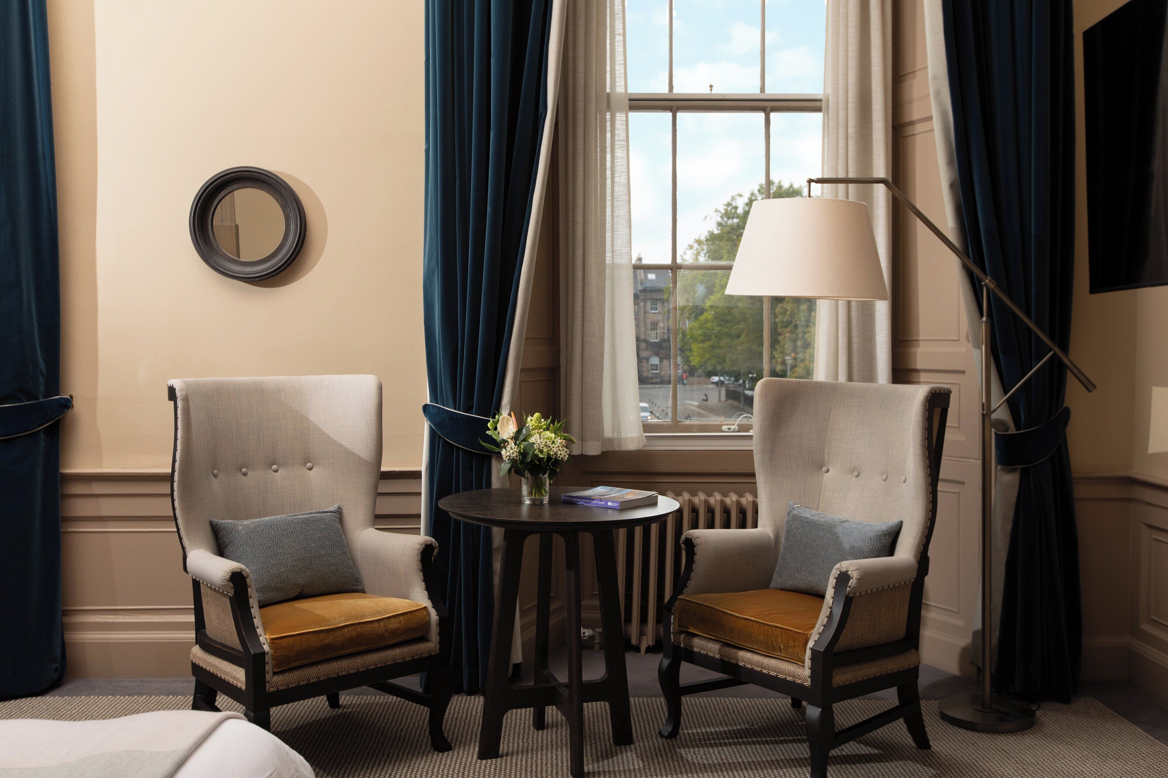 A hotel room with two upholstered armchairs, a small round table with a vase of flowers, a floor lamp, and a view of the city through a window with curtains.
