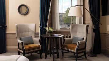 Ein Hotelzimmer mit zwei Polstersesseln, einem kleinen runden Tisch mit Blumenvase, einer Stehlampe und Blick auf die Stadt durch ein mit Vorhängen drapiertes Fenster.