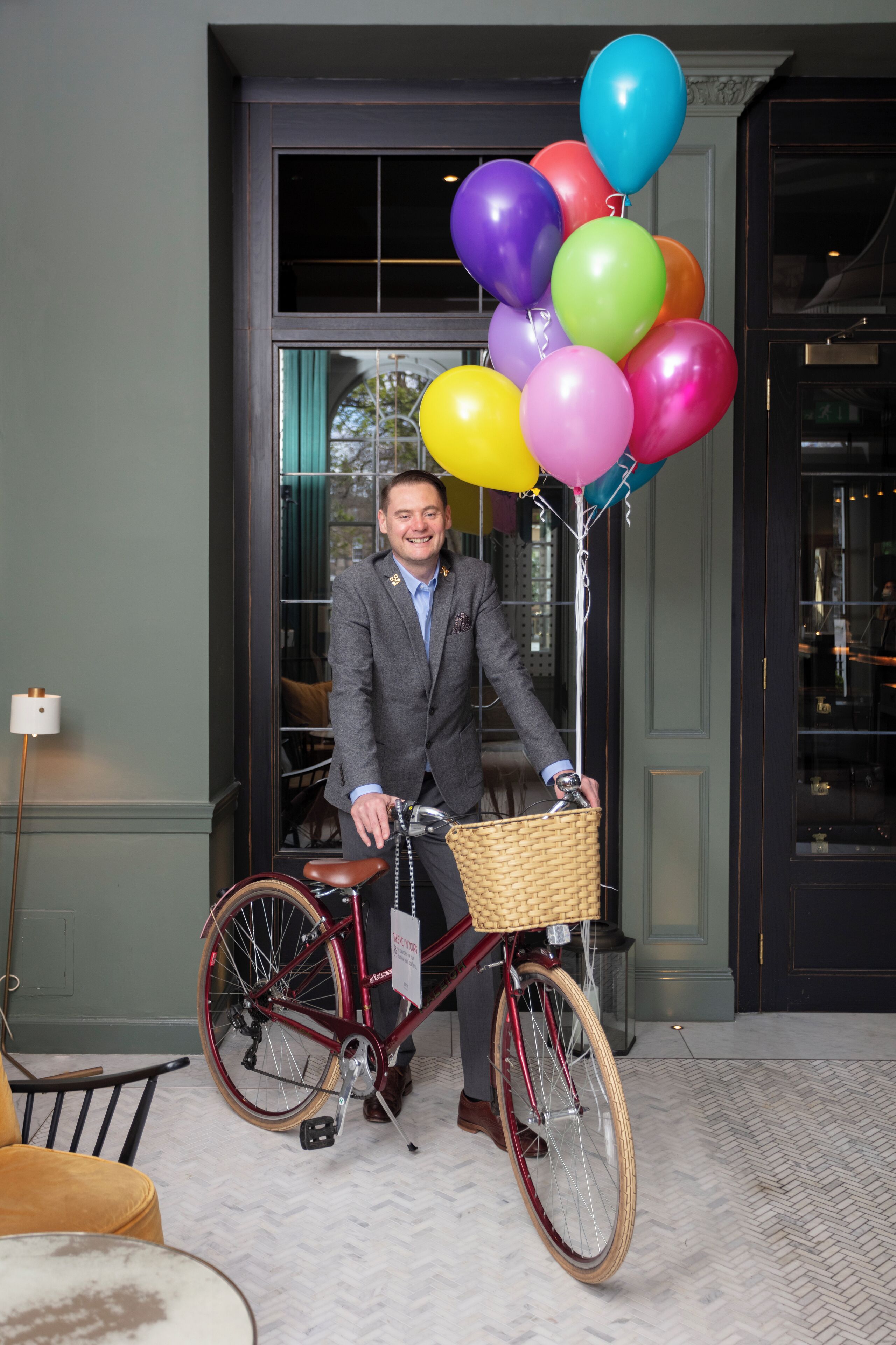 A man in a suit holding a bicycle with a basket and balloons.