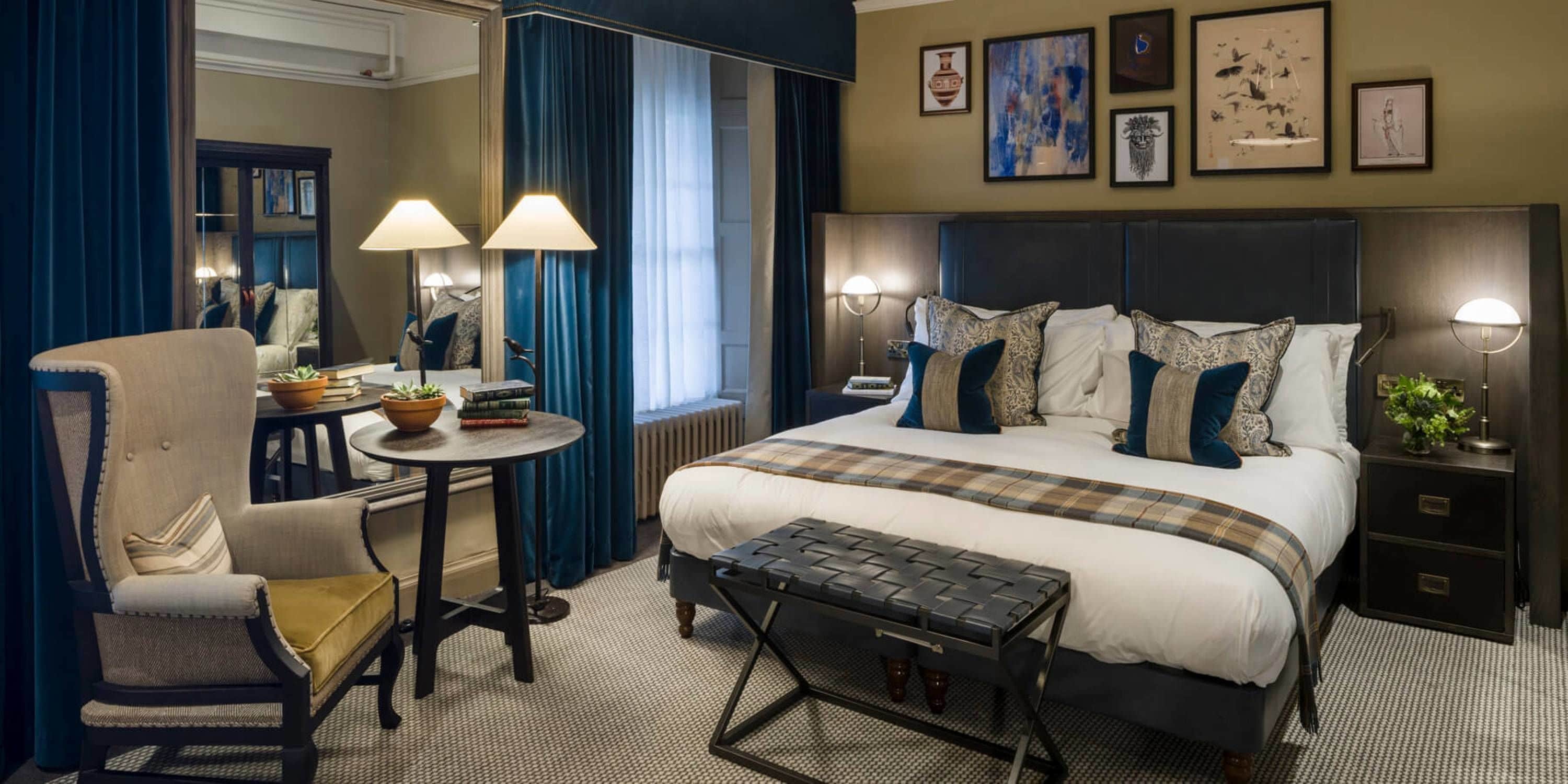A hotel room with a large bed, a chair, a small table, and a bench. There are lamps on either side of the bed and a window with curtains.