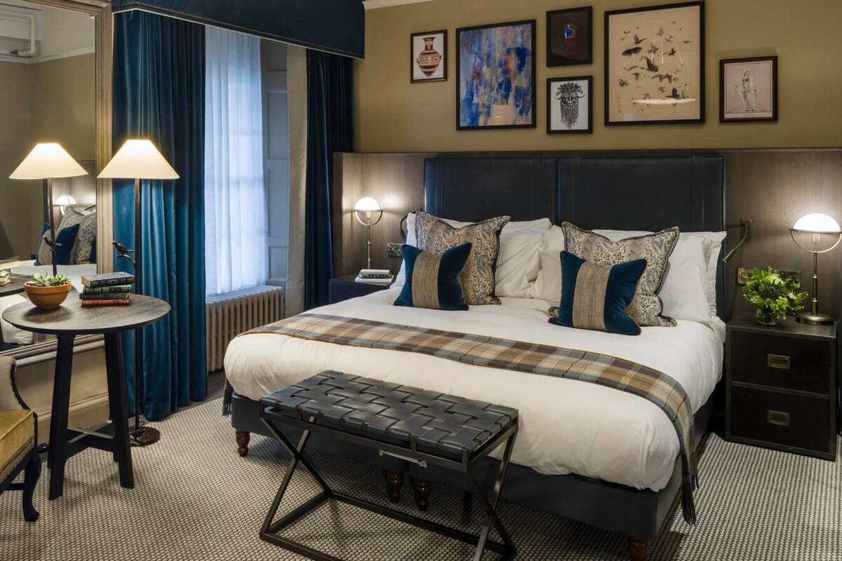 Een hotelkamer met een groot bed, een stoel, een klein tafeltje en een bankje. Er staan lampen aan weerszijden van het bed en er is een raam met gordijnen.