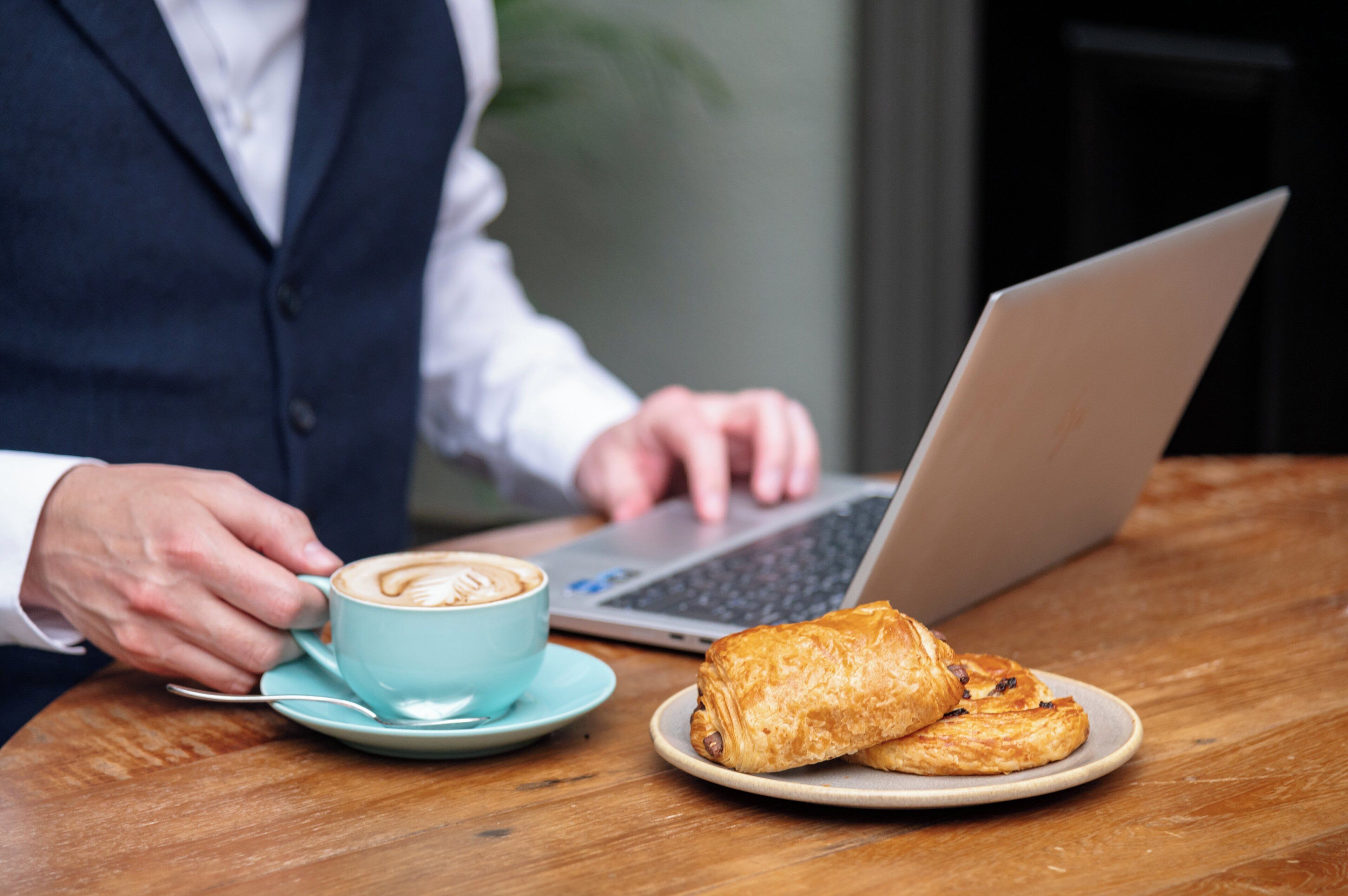 A person in a business suit using a laptop on a wooden table with a cup of coffee and two pastries.