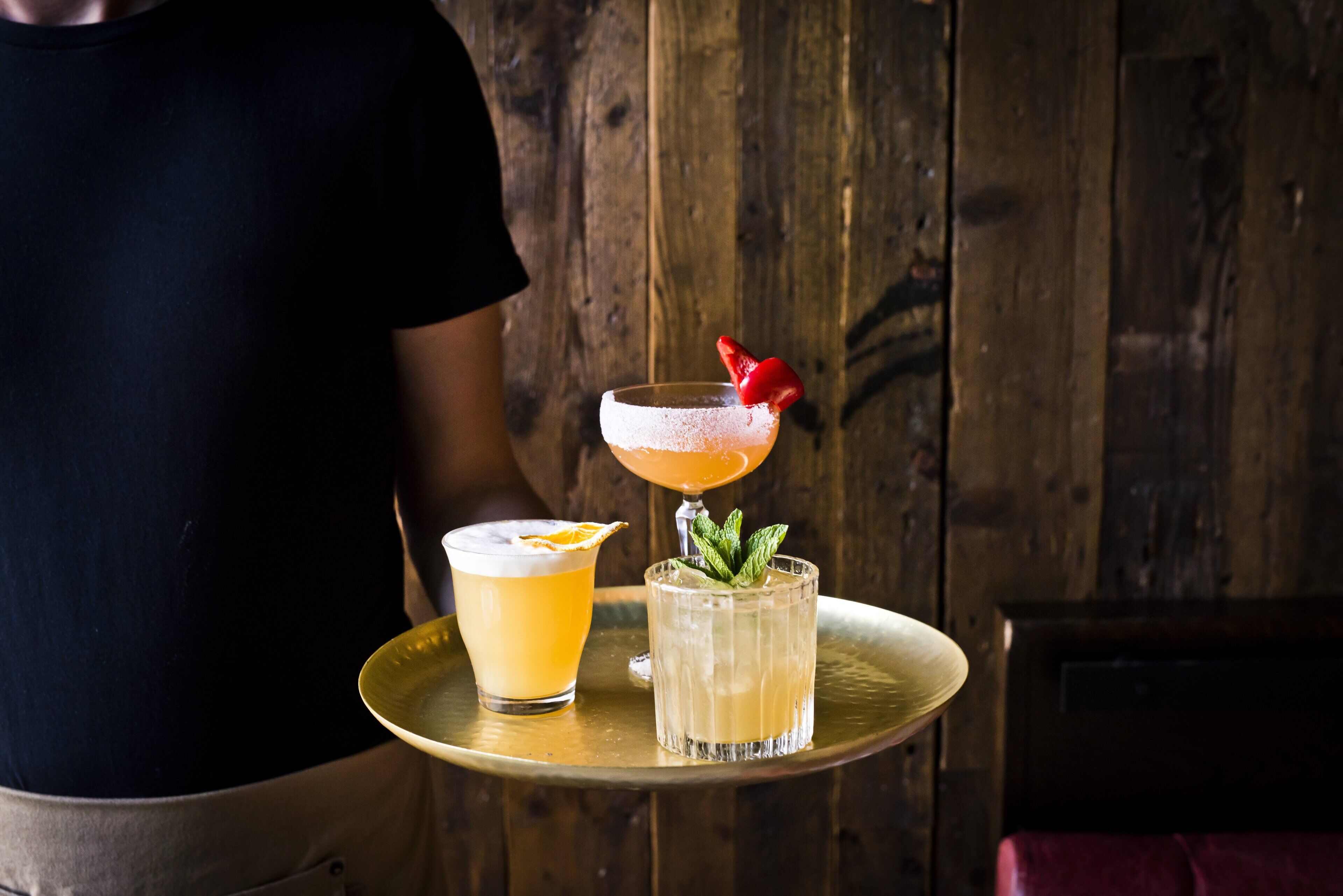 A person holding a tray with two drinks, one with a strawberry garnish and mint leaves, and another with an orange slice garnish.