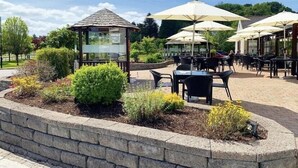 Ein Außenbereich mit einer Terrasse, Steinsitzgelegenheiten, einem Glasgazebo und Sonnenschirmen, die Schatten spenden.