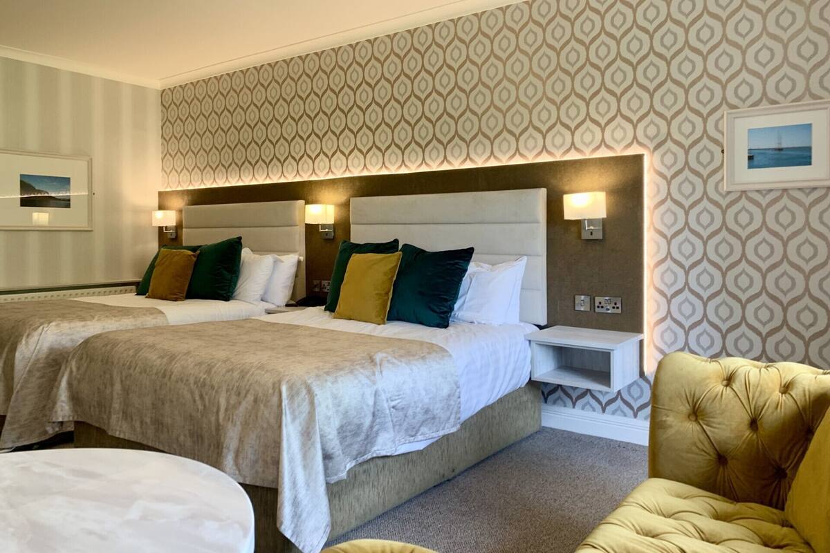 A hotel room with two beds, a sofa, and a chair. There are wall-mounted lamps and a framed picture on the wall.