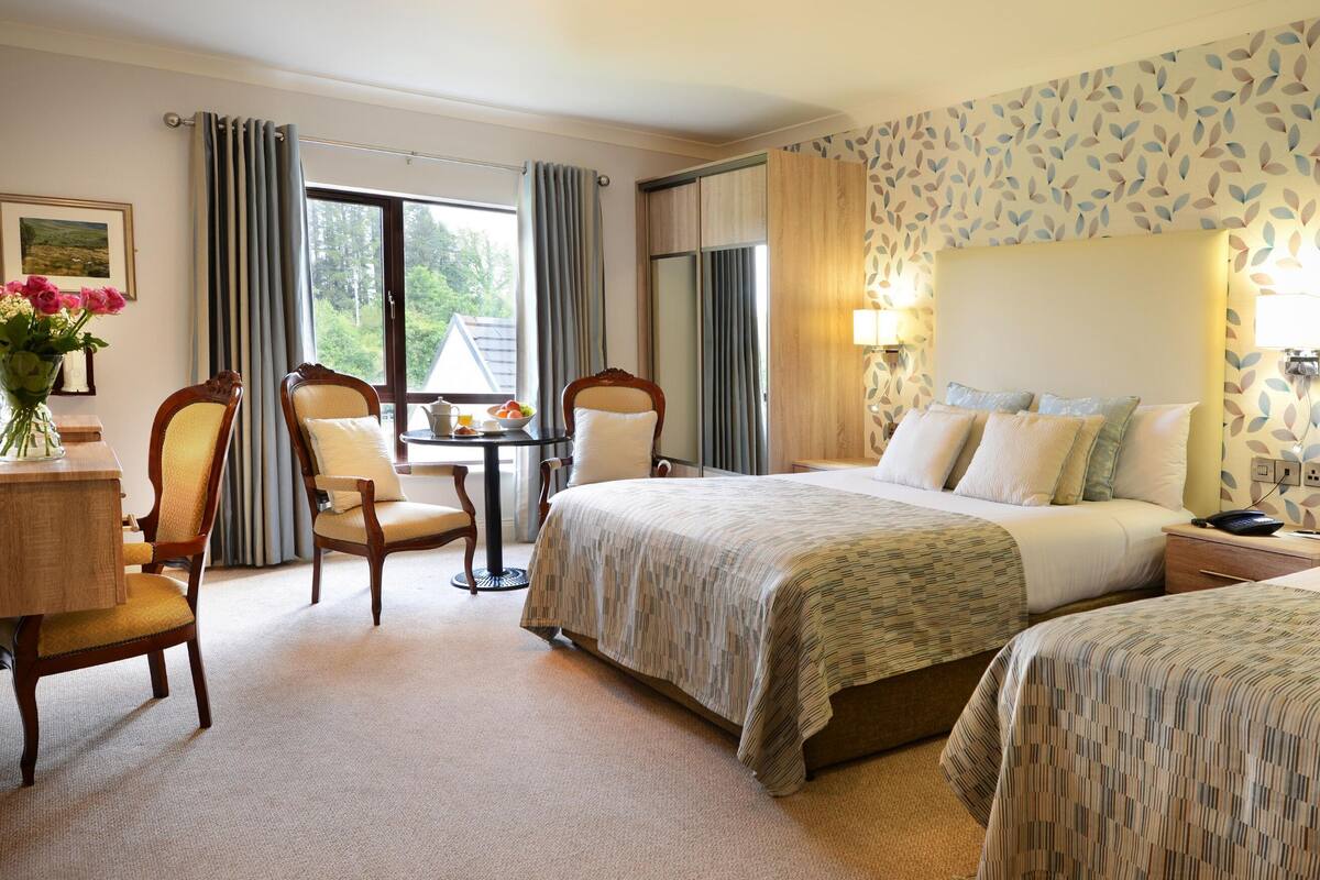 A hotel room with a bed, two chairs, a small table with a vase of flowers, a lamp, and a view of greenery outside.