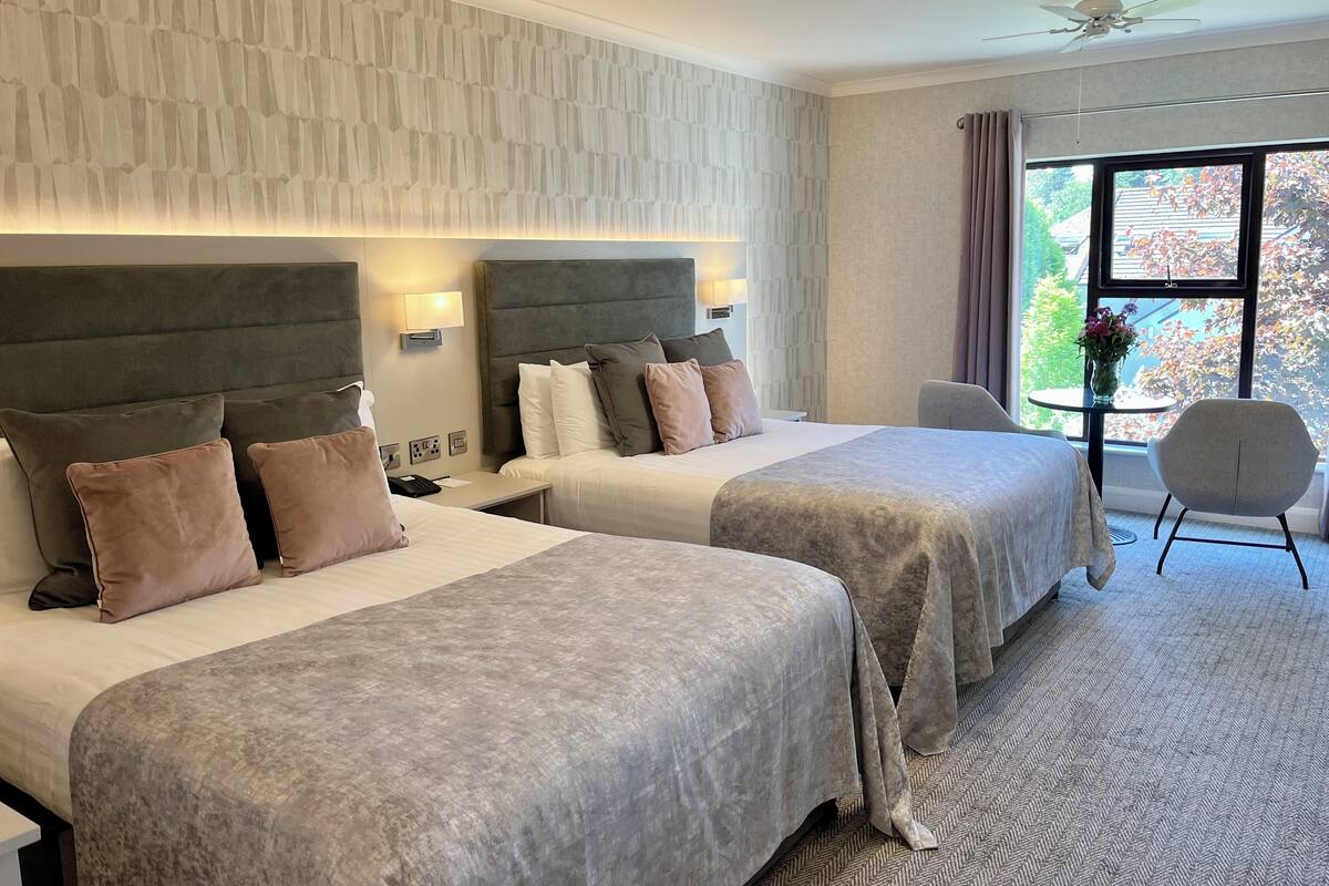 A hotel room with two beds, a small table with a chair, and a large window with a view of trees.