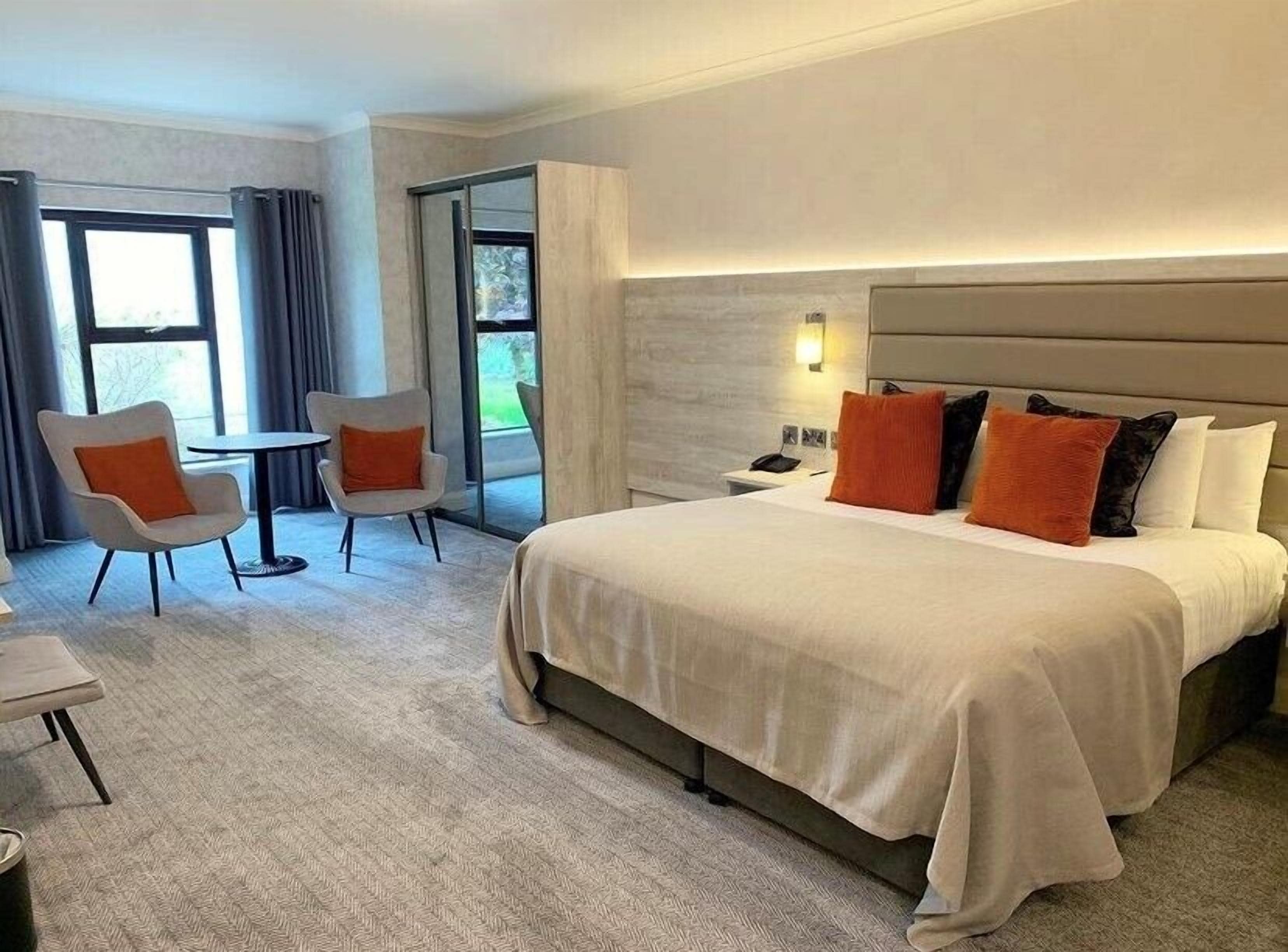 Habitación de hotel con una cama grande, dos sillones, una mesita pequeña y una mesita de noche con teléfono.