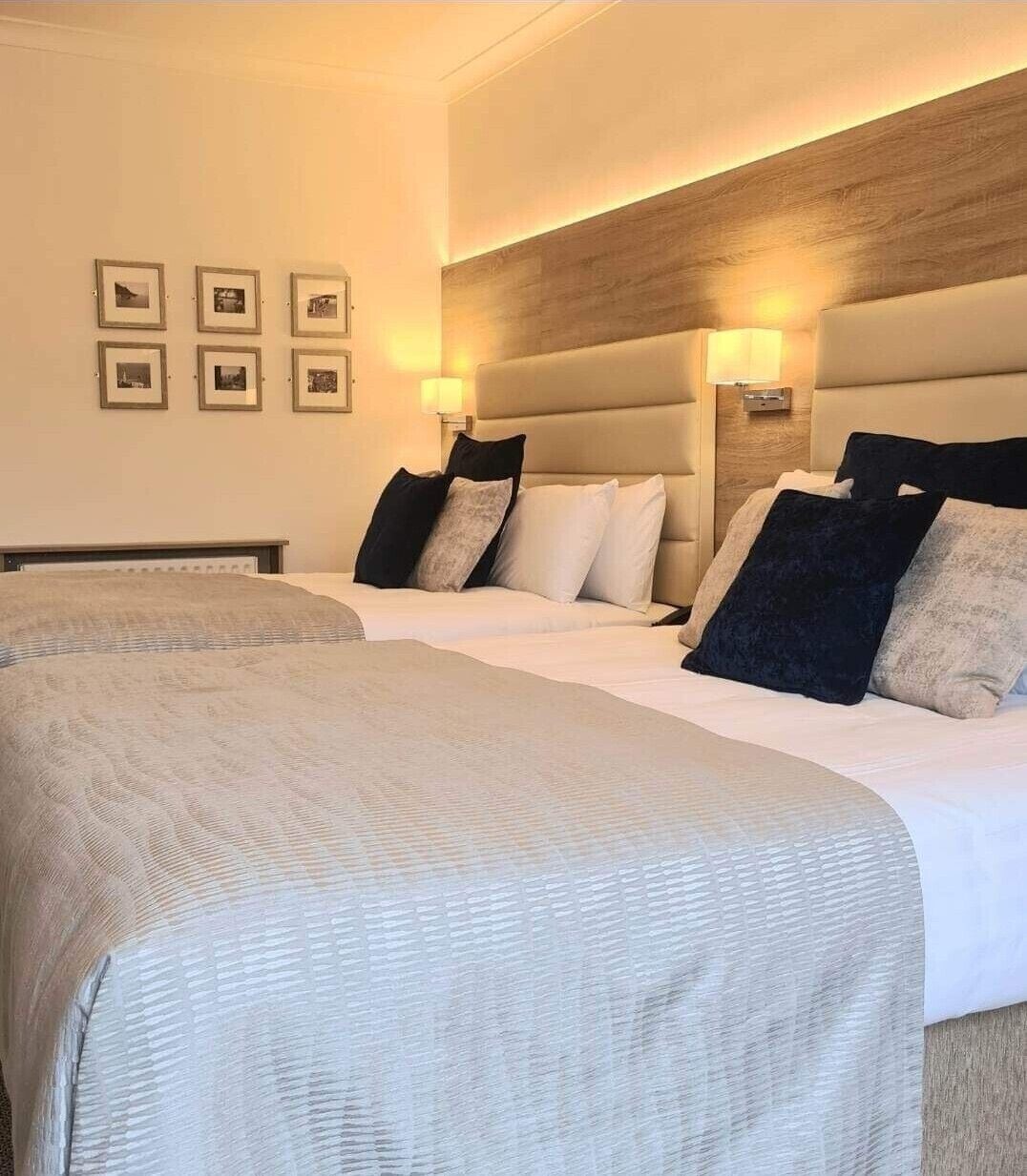 A hotel room with two beds, a wooden headboard, wall-mounted lamps, and framed pictures on the wall.