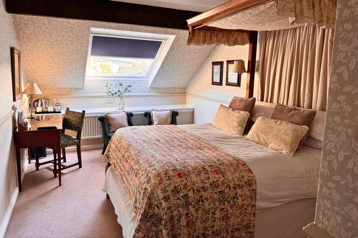 A bedroom with a large bed, a desk with a lamp, a chair, and a window with curtains.