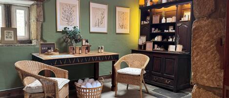 A cozy room with wicker chairs, a wooden table with bottles, framed botanical prints on the wall, and a dark wooden cabinet with shelves.