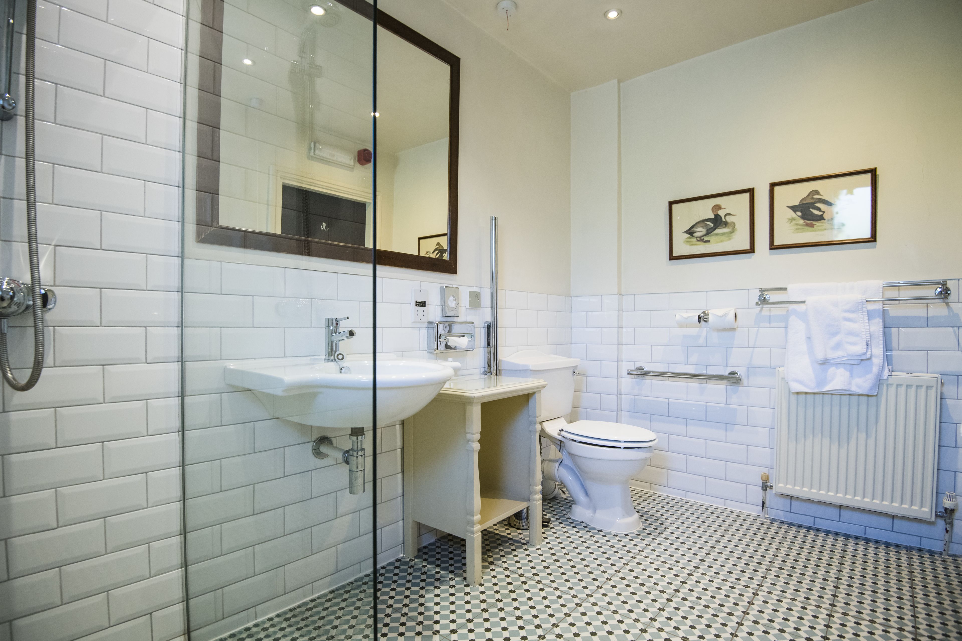 Ein Badezimmer mit weißen Fliesen, einer Glasdusche, einem Waschbecken, einer Toilette und zwei gerahmten Bildern an der Wand.