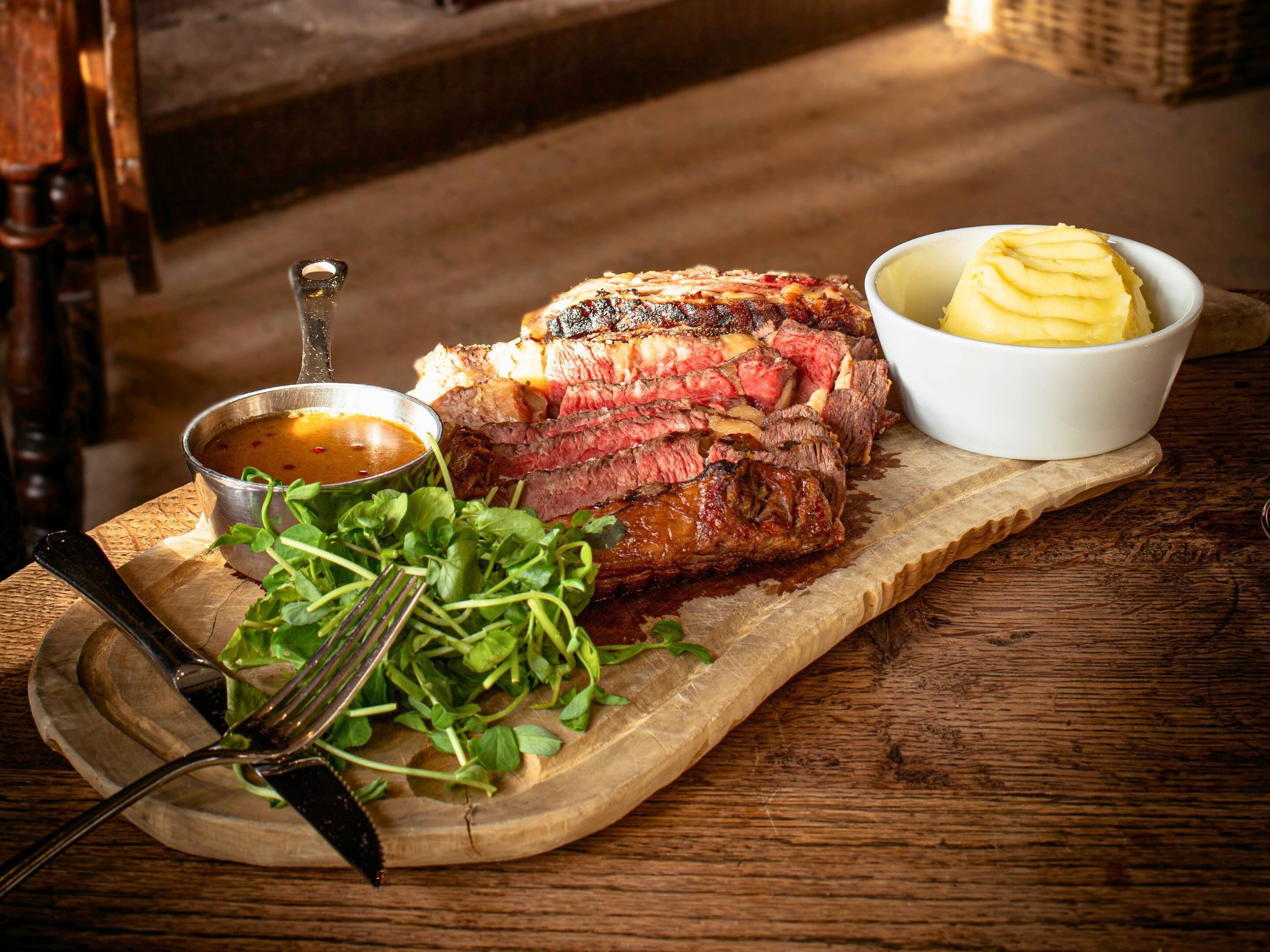 A steak with a side of mashed potatoes and greens on a wooden board.