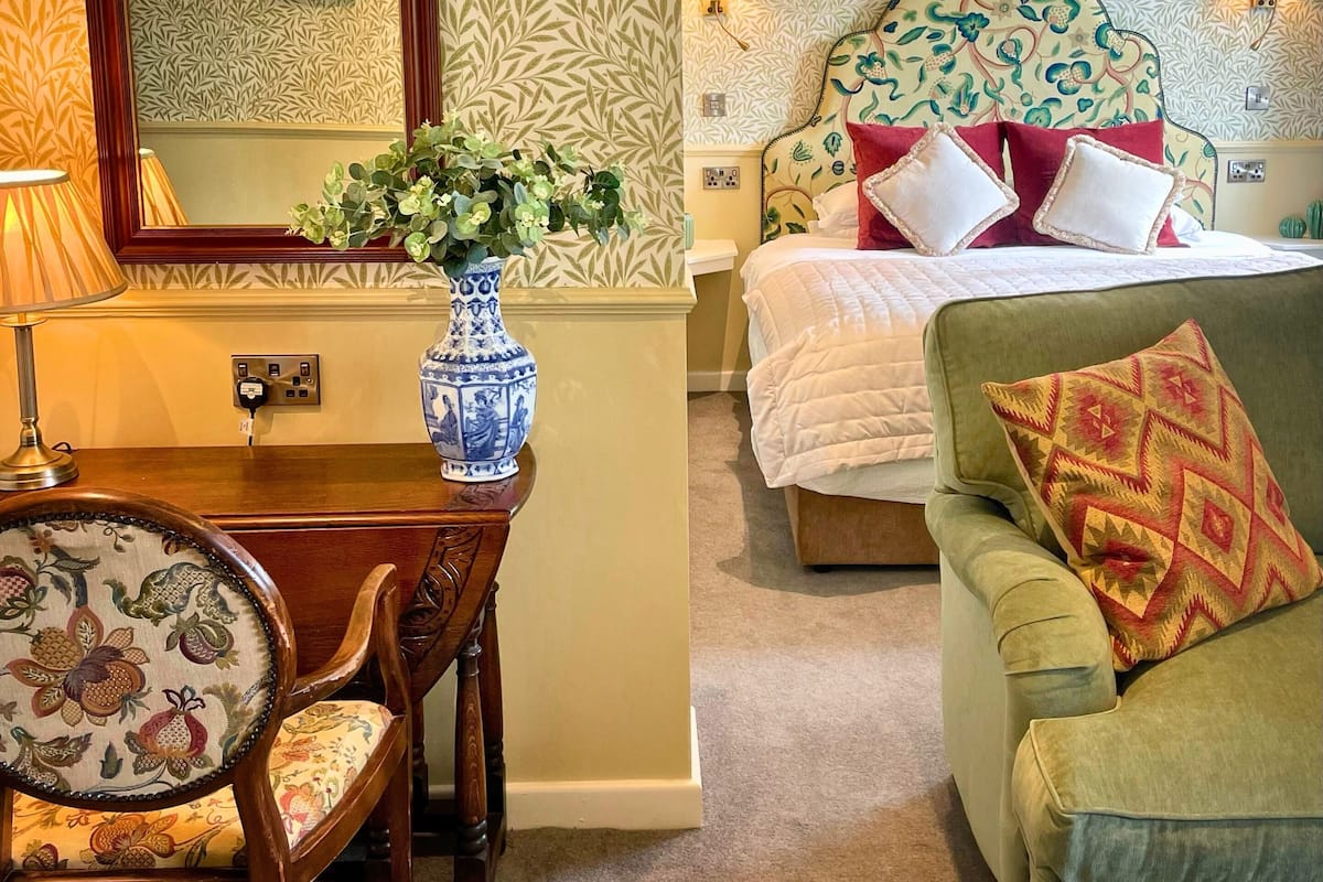 A hotel room with a bed, a green armchair, a wooden table with a vase, and a chair with floral upholstery.