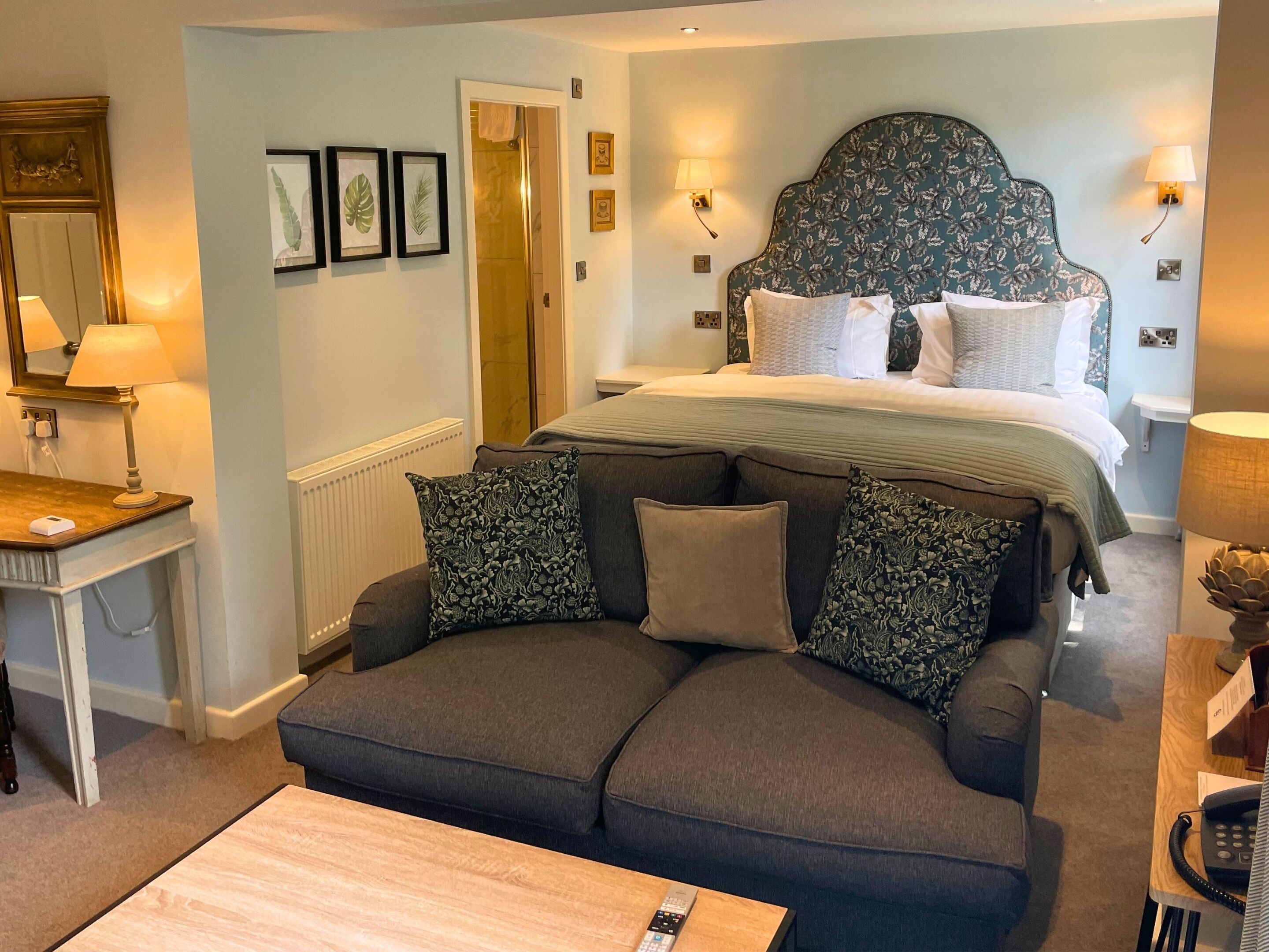 A hotel room with a large bed, a sofa, a desk with a lamp, and framed pictures on the wall.