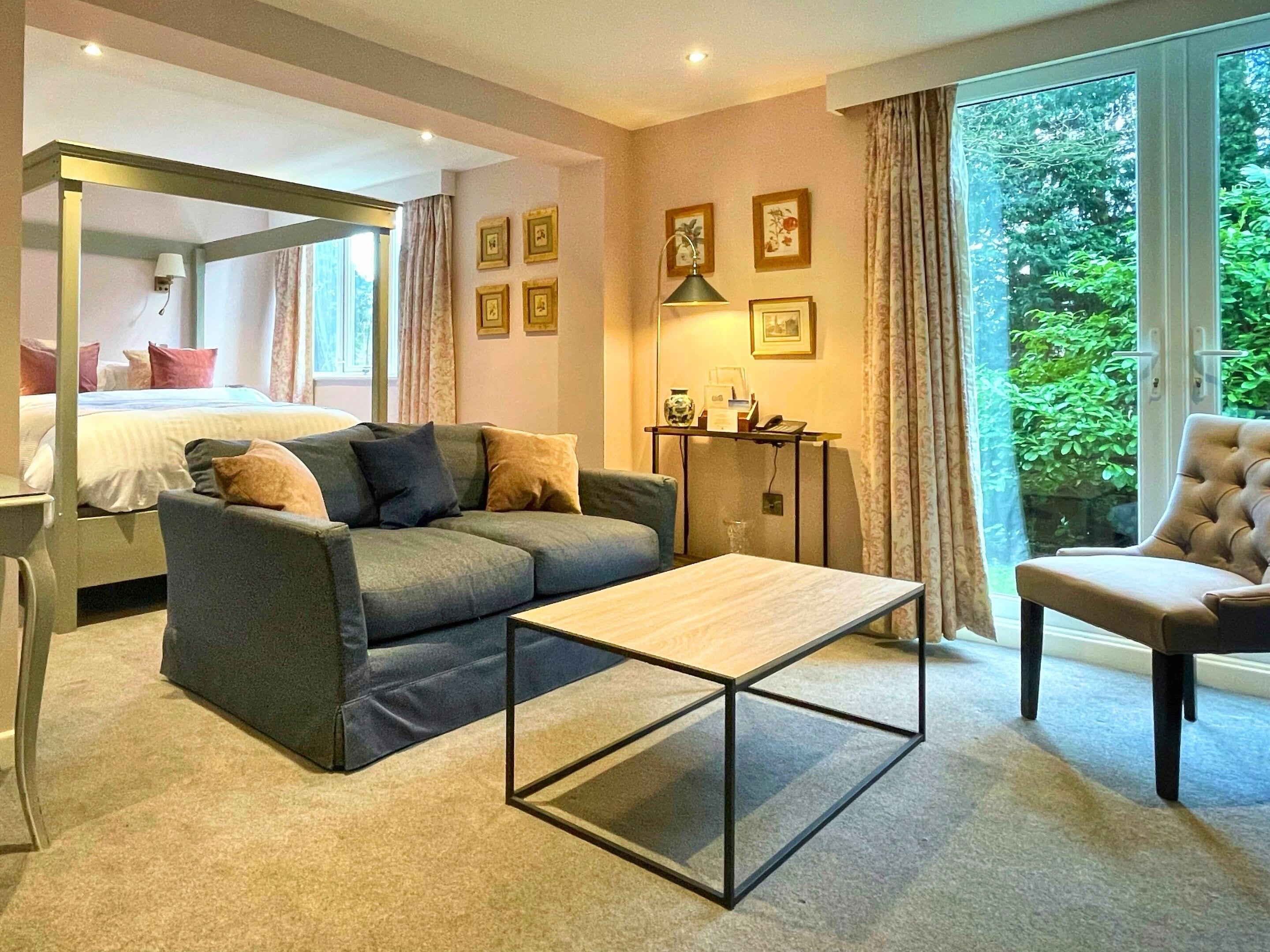 Une chambre d’hôtel moderne, équipée d’un lit, d’un canapé, d’une table basse et d’un fauteuil. Des tableaux encadrés ornent le mur, et une fenêtre offre une vue sur la verdure.