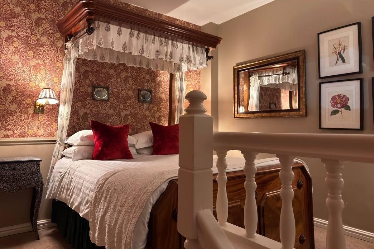 A four-poster canopy bed with a white bedspread and red pillows, a wooden nightstand with a lamp, and a mirror on the wall.