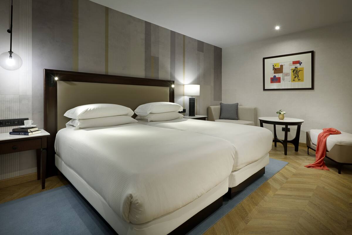 A hotel room with a large bed, bedside tables, a desk, a chair, and a small table with a lamp.