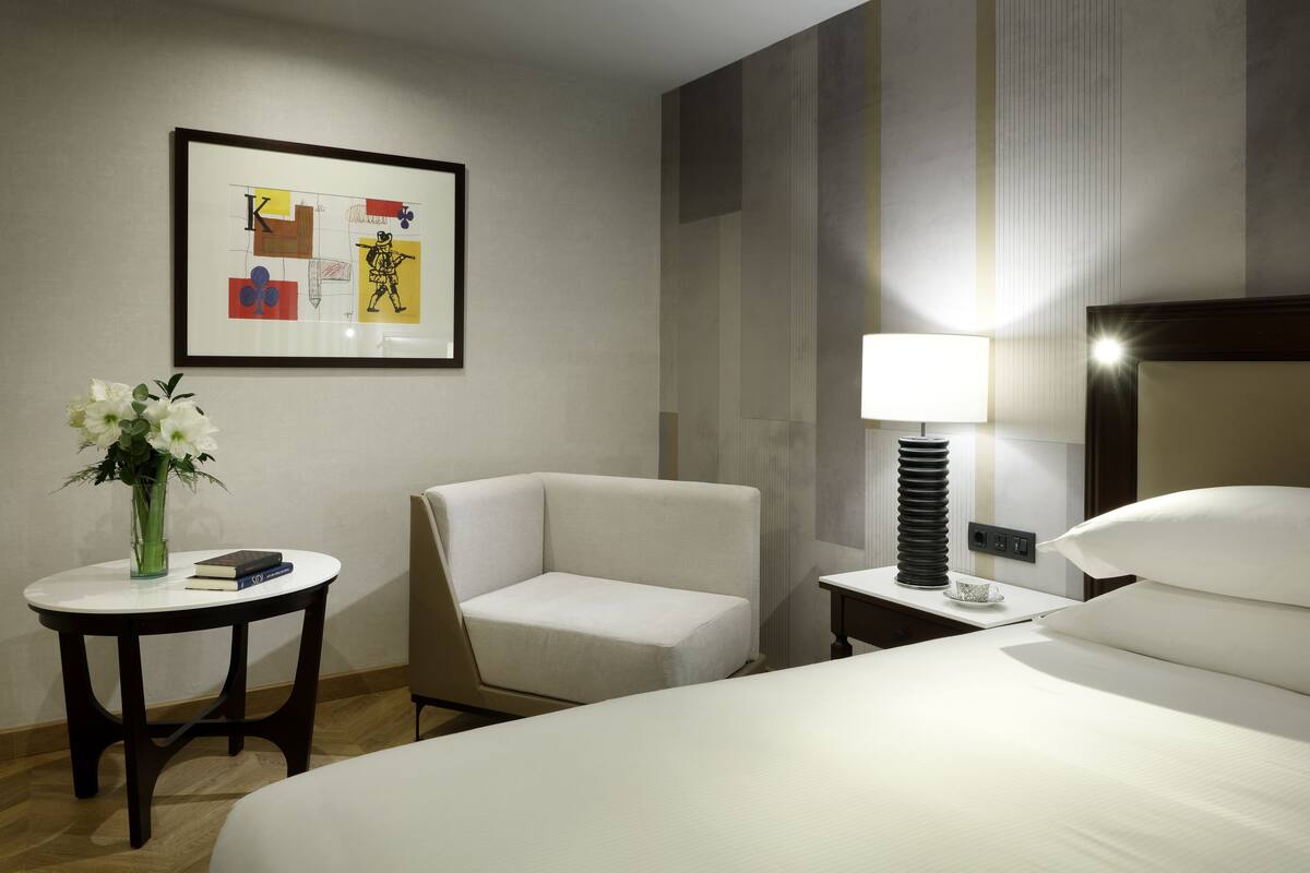 A hotel room with a bed, a chair, a small table with a vase and book, a nightstand with a lamp, and a framed artwork on the wall.